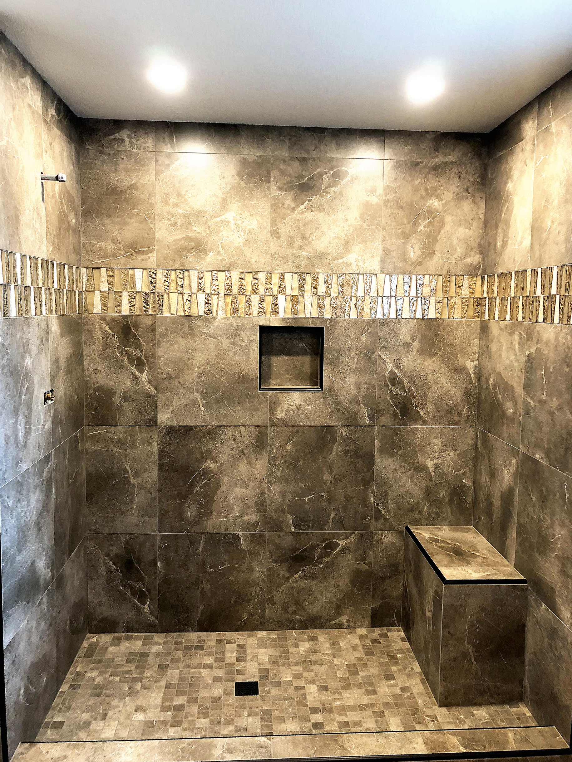 A modern walk-in shower with brown marble tiles, a decorative horizontal strip of smaller tiles, a built-in niche, a small bench, and a gray floor drain.