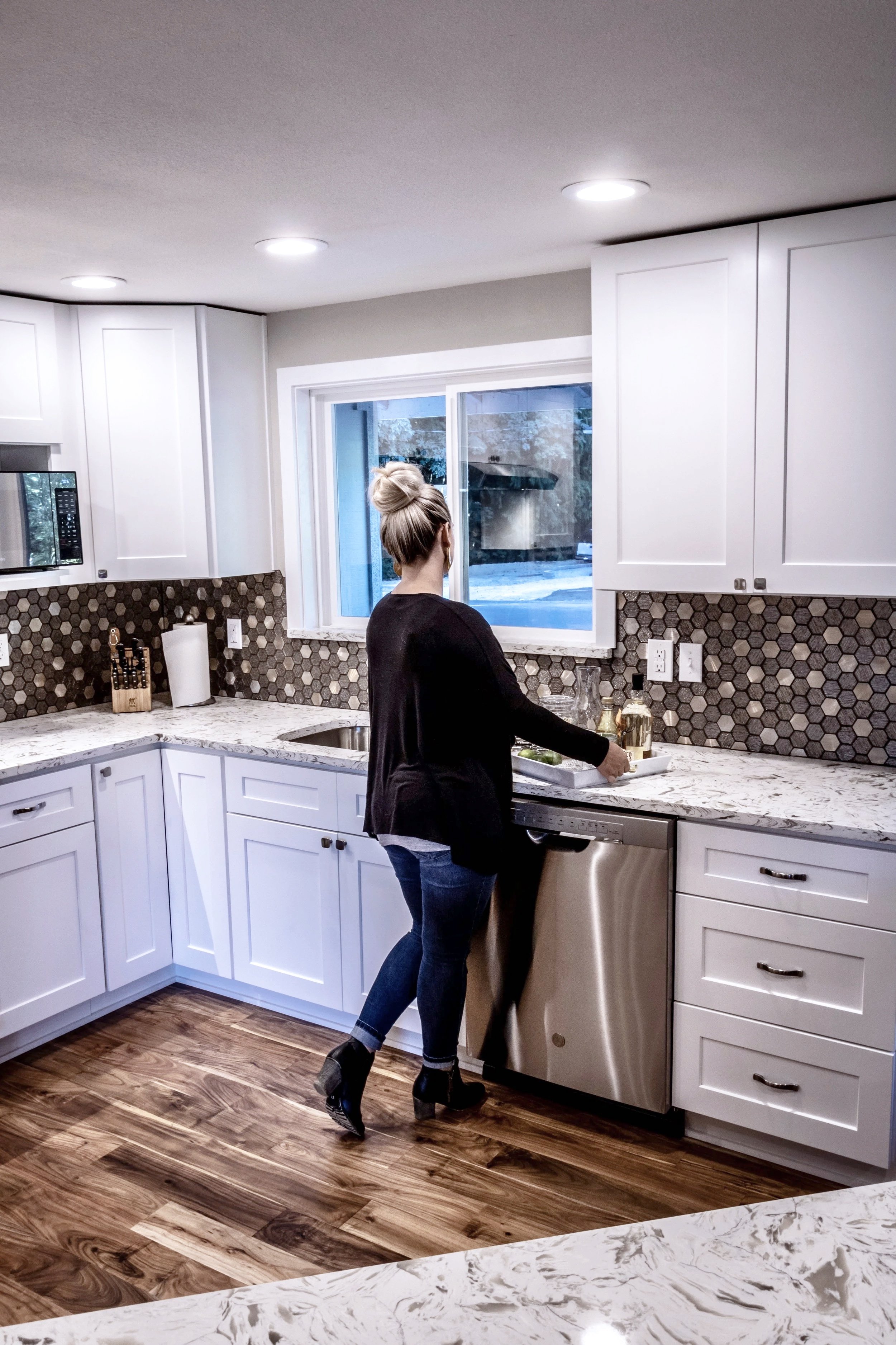 A woman standing in a modern kitchen near the sink, dressed in a black top, jeans, and black heeled boots, looking out the window.