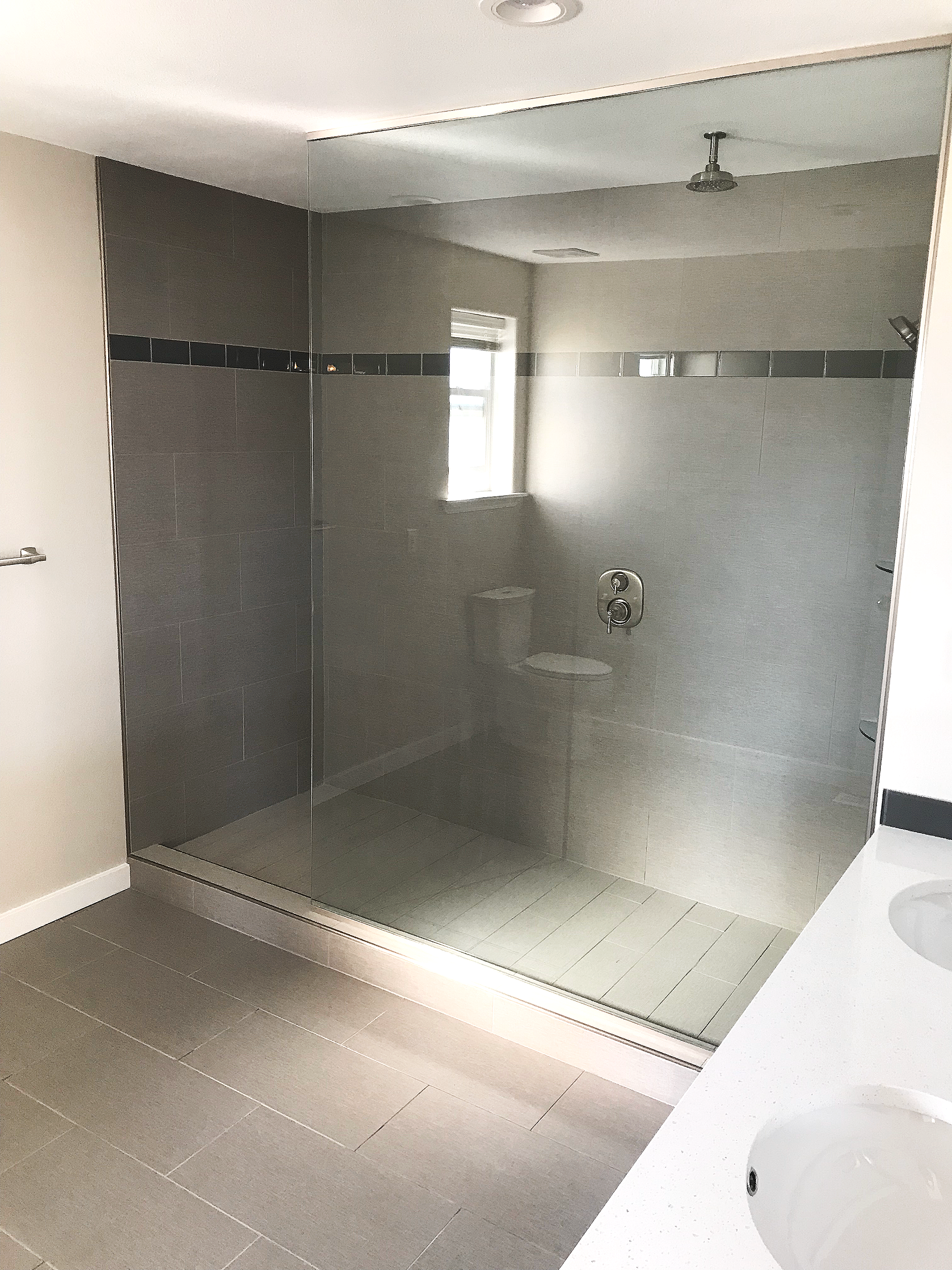 Modern bathroom with walk-in glass shower featuring gray tiles and a black accent strip, a white countertop with two sinks, a small window, and neutral wall colors.