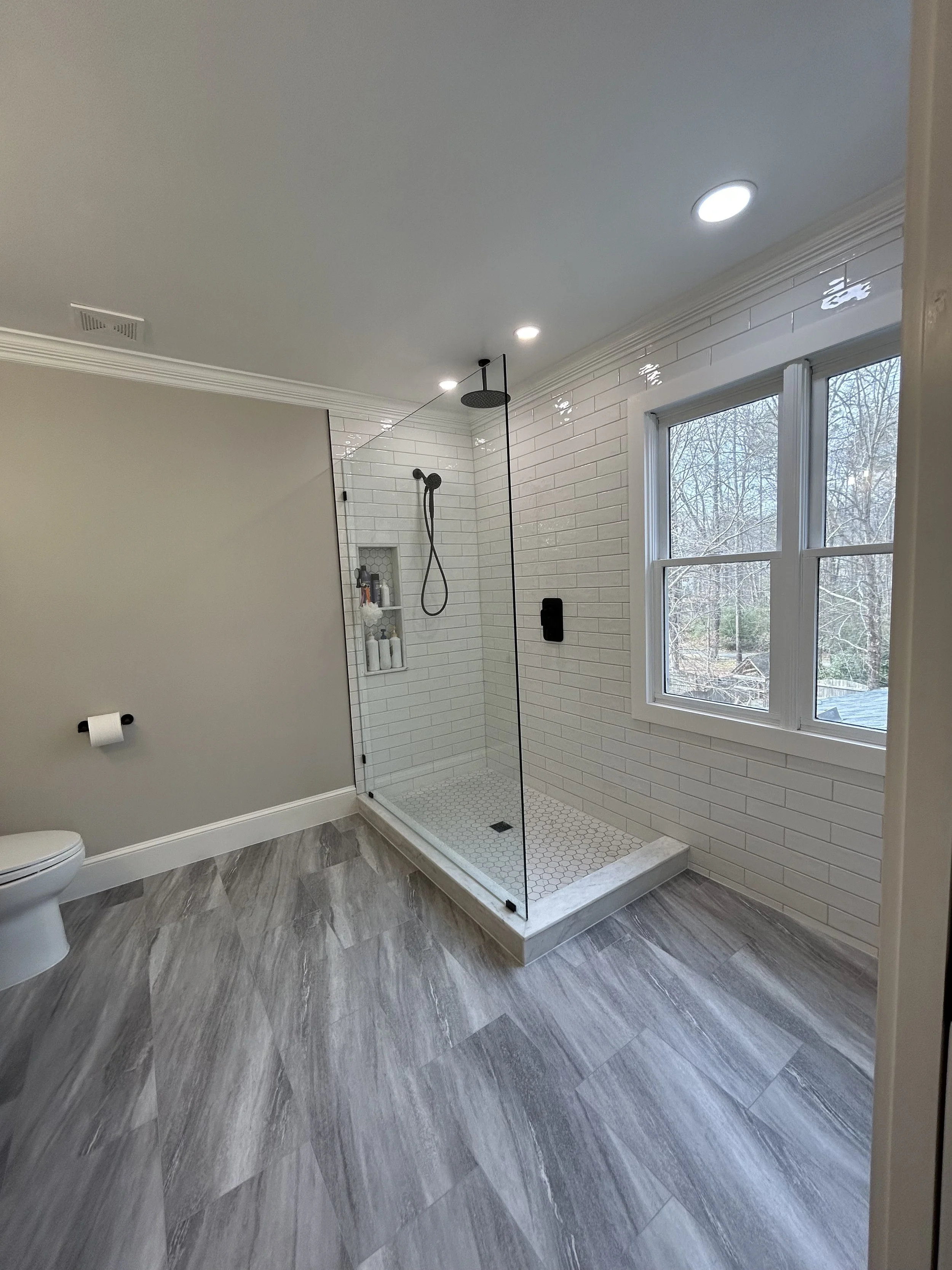 A modern bathroom with a walk-in shower enclosed by a glass panel, white subway tile walls, a large window, and gray wood-like floor tiles. There is a toilet with a mounted toilet paper holder on the wall.