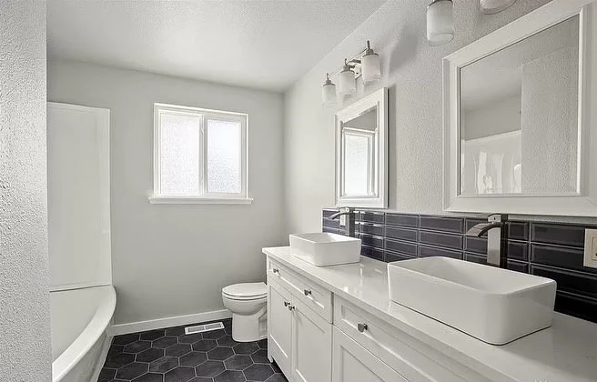 A modern bathroom with white cabinetry, black hexagonal floor tiles, a bathtub, a toilet, and two white vessel sinks with mirrors above, illuminated by three wall-mounted lights.