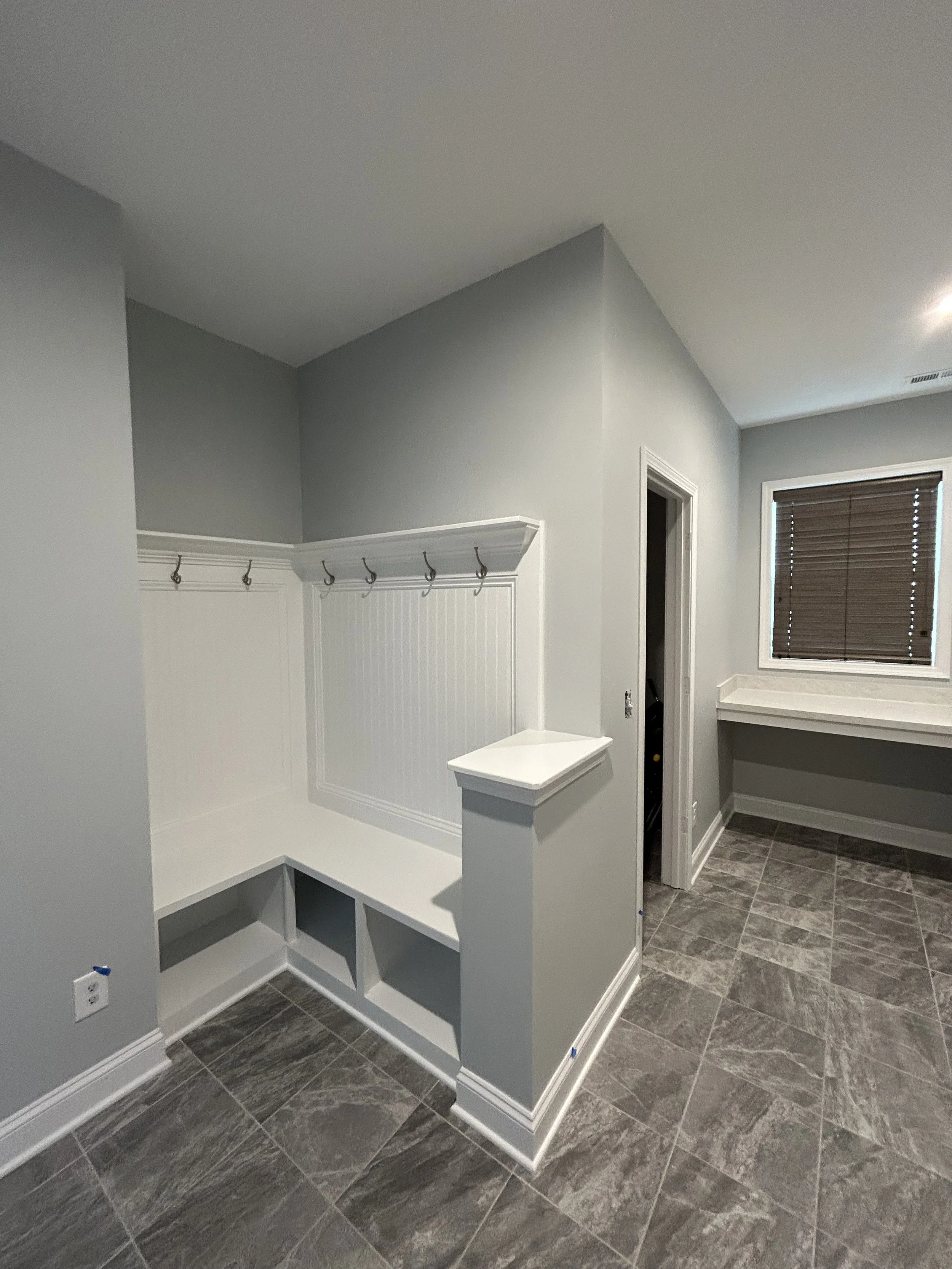 Empty laundry room with gray tile floors, white wainscoting, coat hooks, built-in bench, and a window with brown blinds.