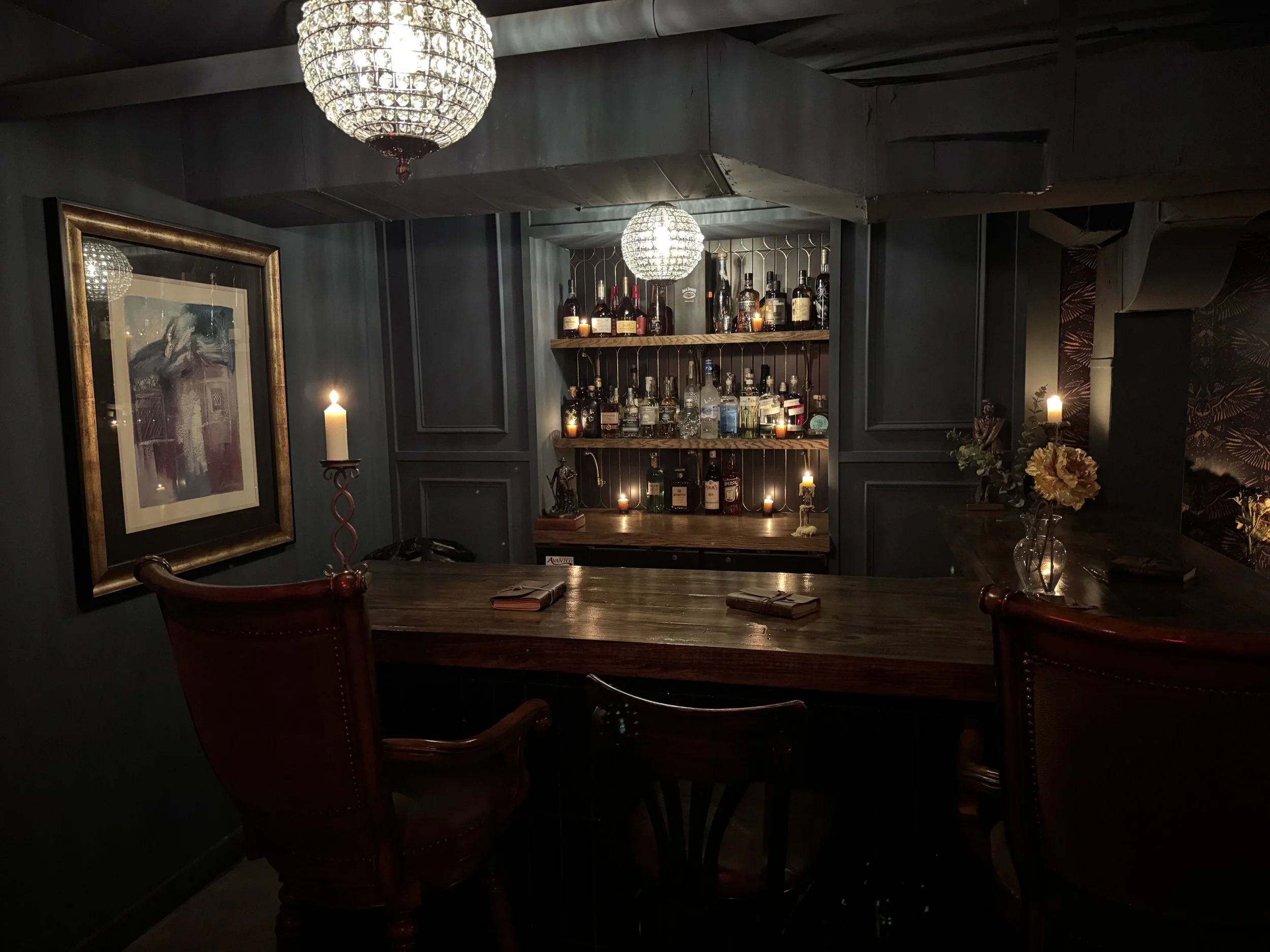 A cozy dimly lit bar with a dark blue wall, two chandeliers, a framed painting, candles, a wooden bar with bottles, and a flower vase on the counter.