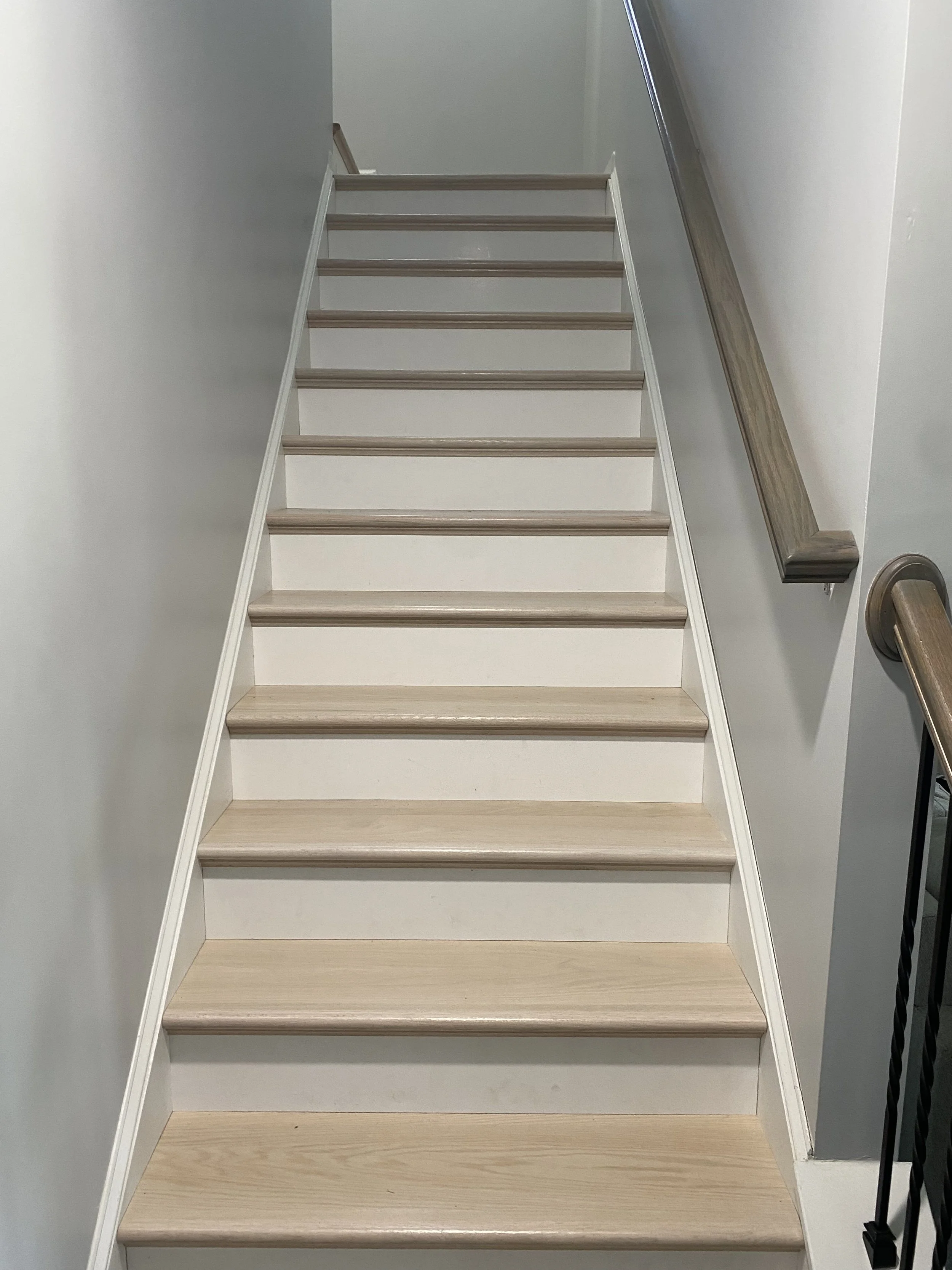 Indoor staircase with light wood steps and white risers, beige baseboards, and a dark wood handrail on the right side mounted on a white wall.