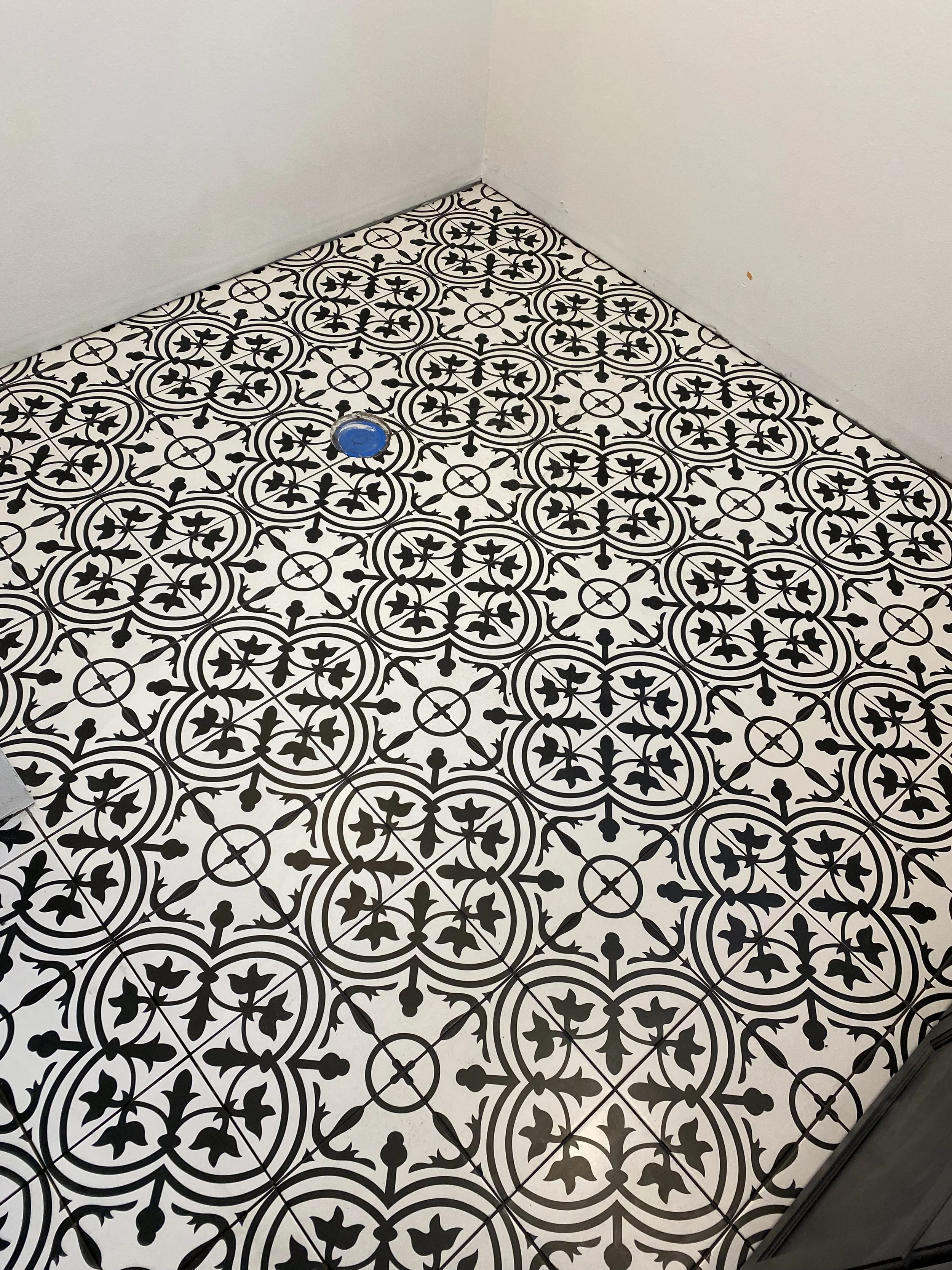 Decorative black and white patterned tile flooring in a corner of a room with a white wall and a blue capped pipe in the middle of the floor.