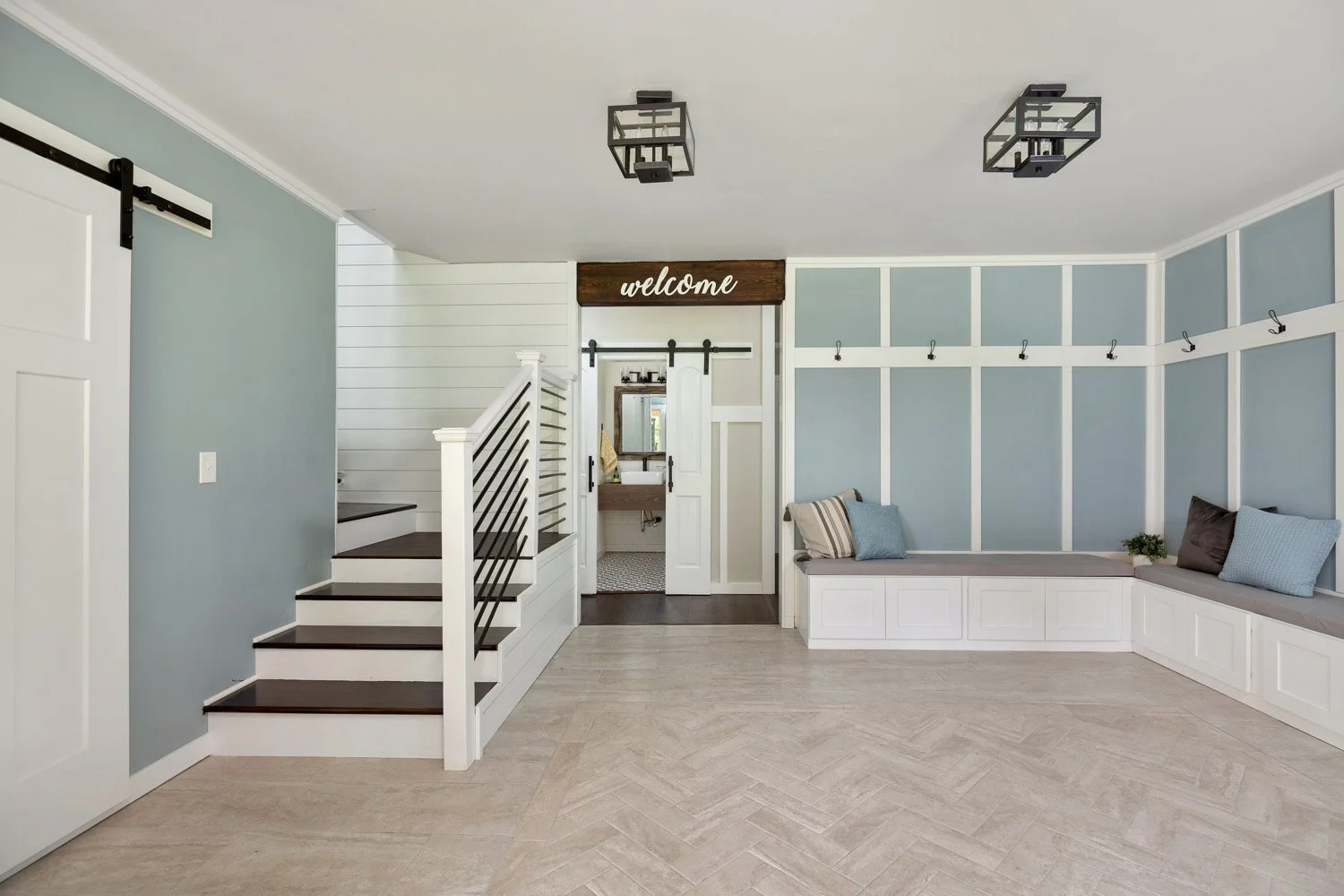 Interior view of a modern home entryway with light blue walls, white trim, and a built-in window seat with pillows, a staircase with black railings, and a wooden 'welcome' sign above a door leading to a bathroom.