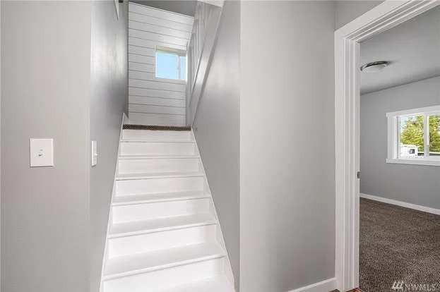 Indoor staircase with white steps and a small window at the top, leading to an upper level, with light gray walls and a doorway to a room with beige carpet and a window.