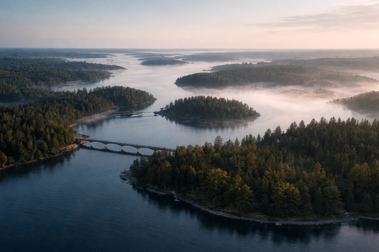 Aerial view of a large body of water with numerous islands covered in dense green trees, a bridge crossing part of the water, and mist or fog over the water and islands during sunrise or sunset.