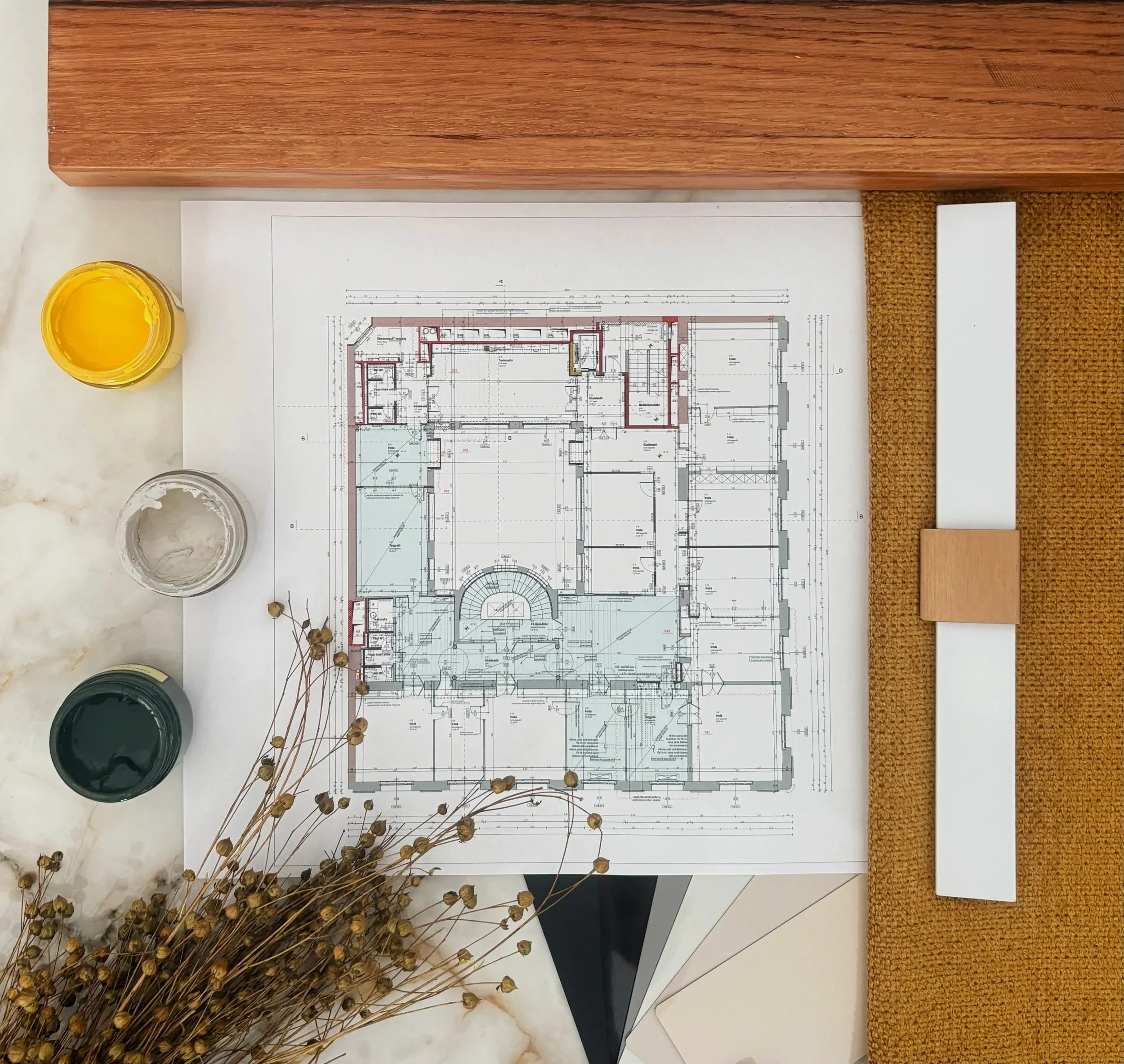Architectural blueprints on a white table with jars of paint, dried flowers, a wooden block, and a brown textured mat nearby.