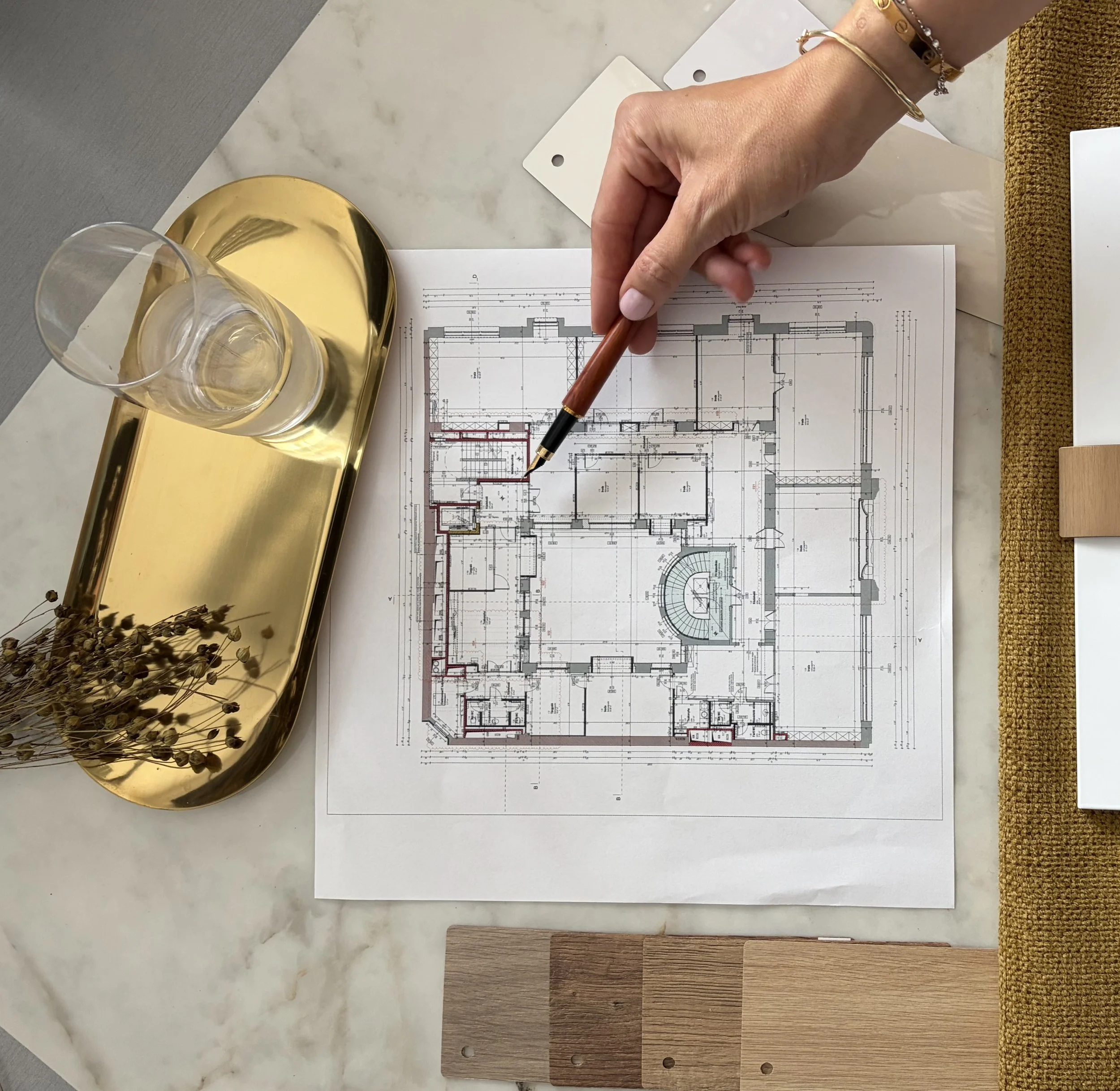 Architectural floor plans on paper, alongside samples of wood and laminate flooring, with a gold tray holding two glasses of water, on a marble tabletop.