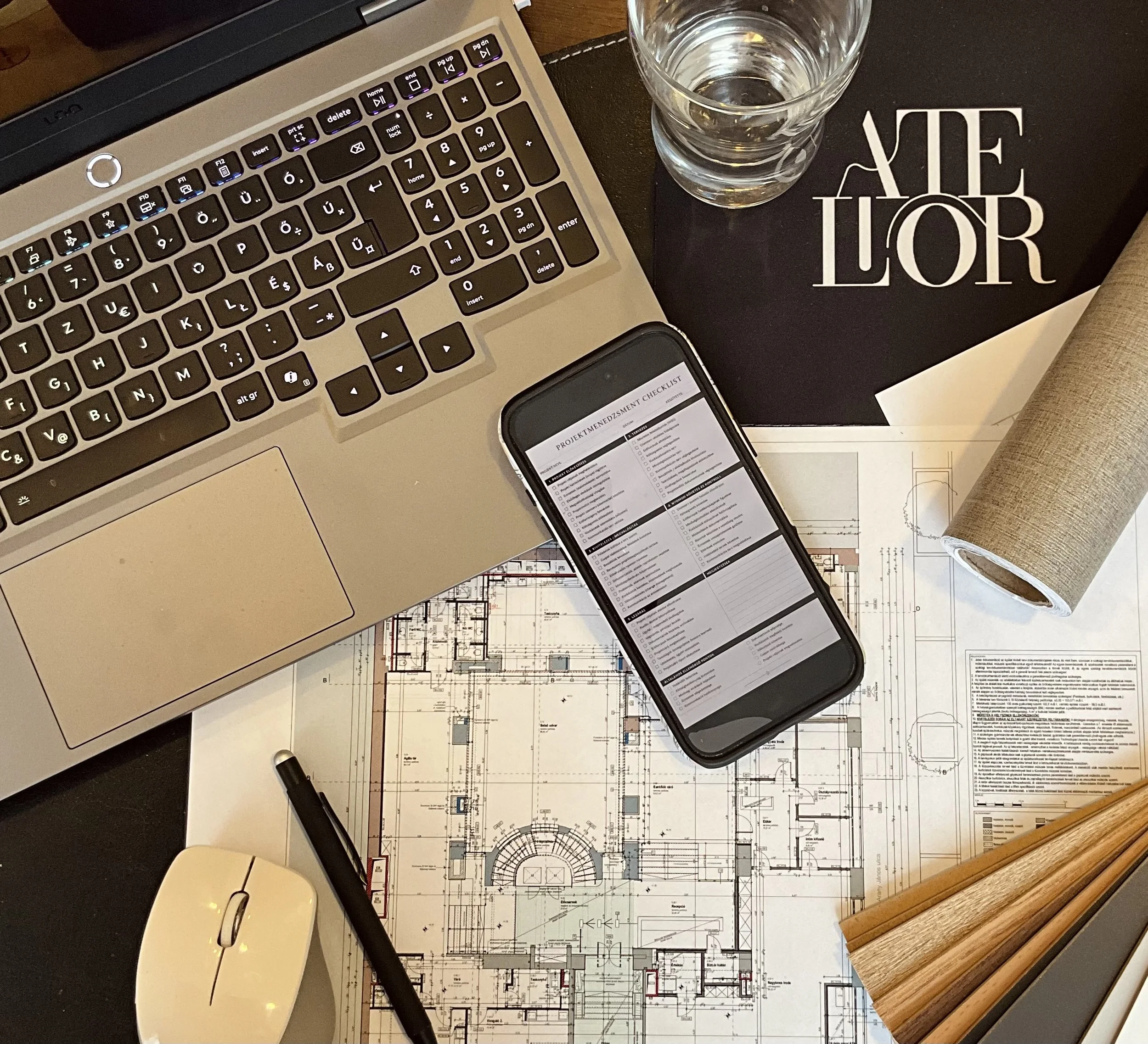 Work desk with a silver laptop showing a black keyboard, a glass of water, a smartphone with a checklist, architectural blueprints, a notebook with white text, a black pen, and a wooden ruler.