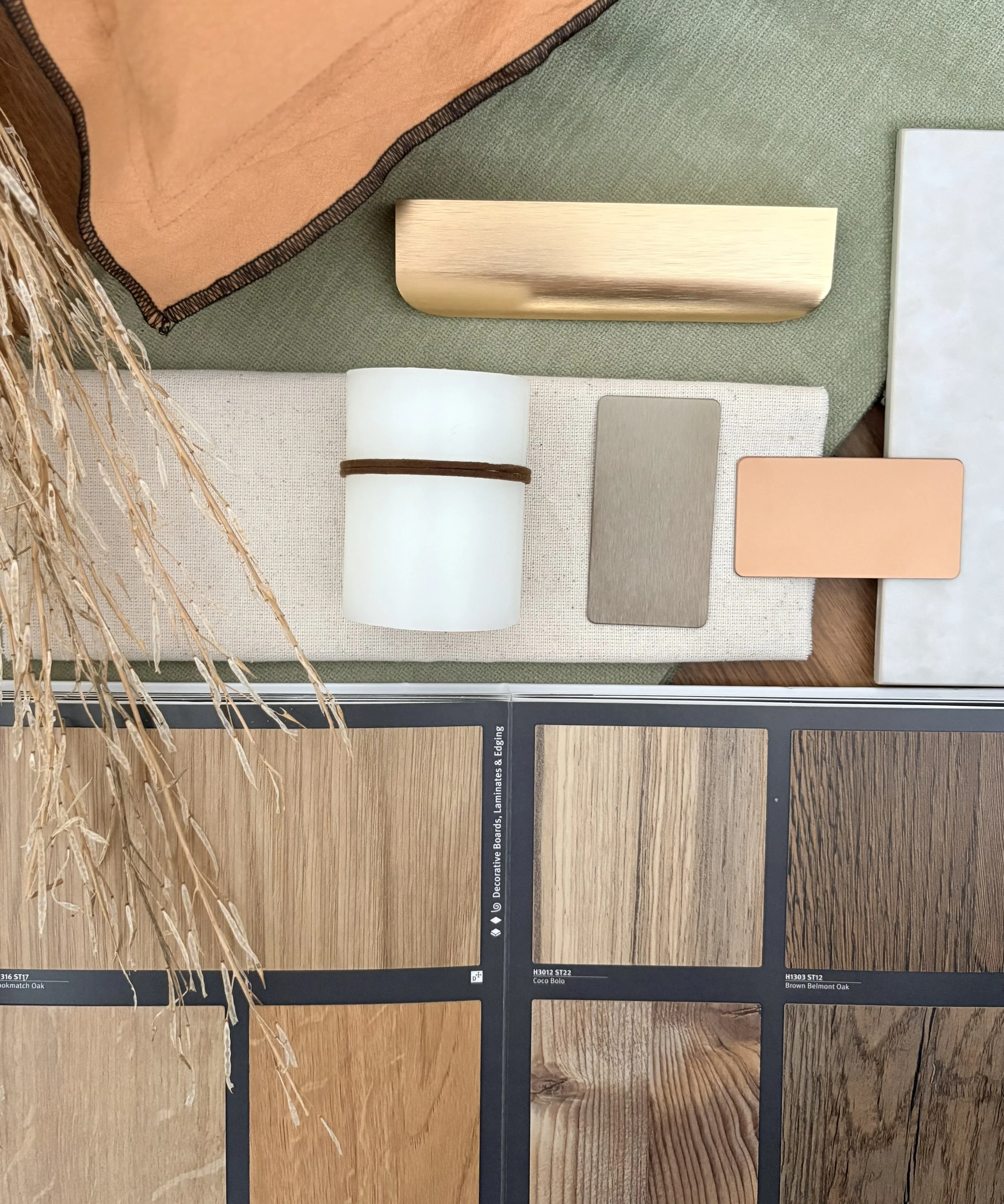 Display of four wood laminate samples, including Kemaratch Oak, Coco Bolo, and Brown Belmont Oak, with a fabric swatch, a small rectangular sample, and a rolled-up white material, arranged on a surface with some decorative dried grass on the left side.
