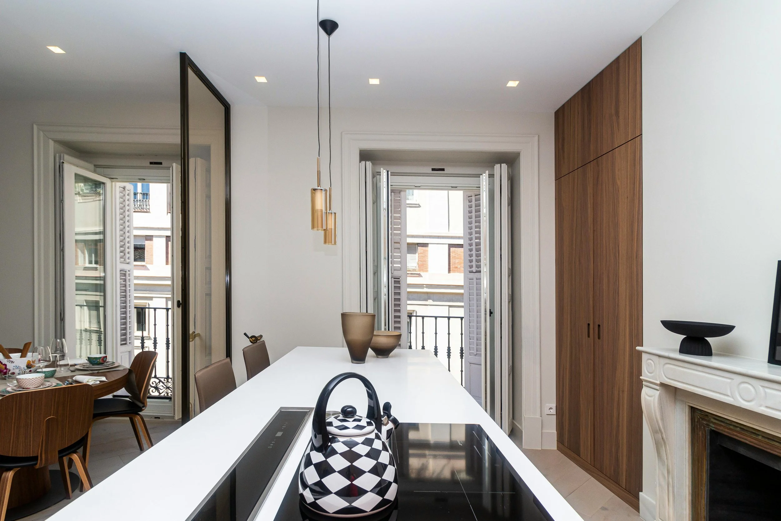 Modern kitchen with white countertops, black and white patterned teapot, vases on the counter, and open double doors leading to a balcony with city buildings in the background.