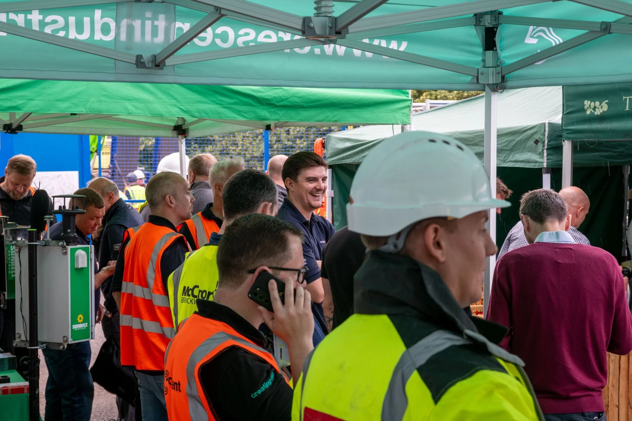 A group of people, including construction workers wearing bright orange and yellow safety vests, some with branded logos, are gathered under green tents at an outdoor event. The group includes a man on the phone, a smiling man in a dark shirt, and a man in a white safety helmet. Other people are standing around, some looking at displays or talking. The background features a fence, trees, and construction equipment.