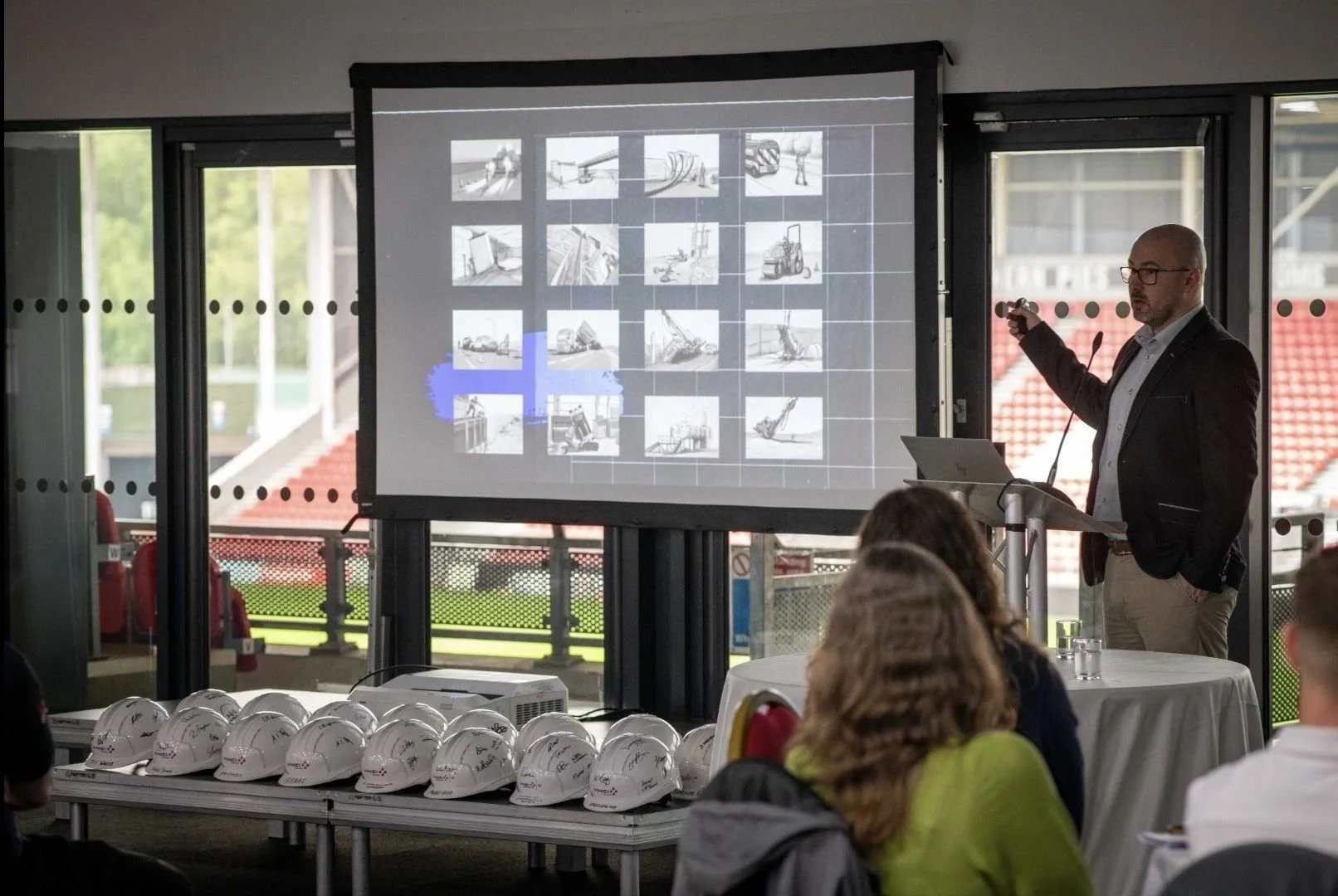 A man in glasses and a black blazer giving a presentation indoors, pointing at a projected slide showing construction equipment images, with several white construction helmets on a table in front of him and an audience seated nearby.