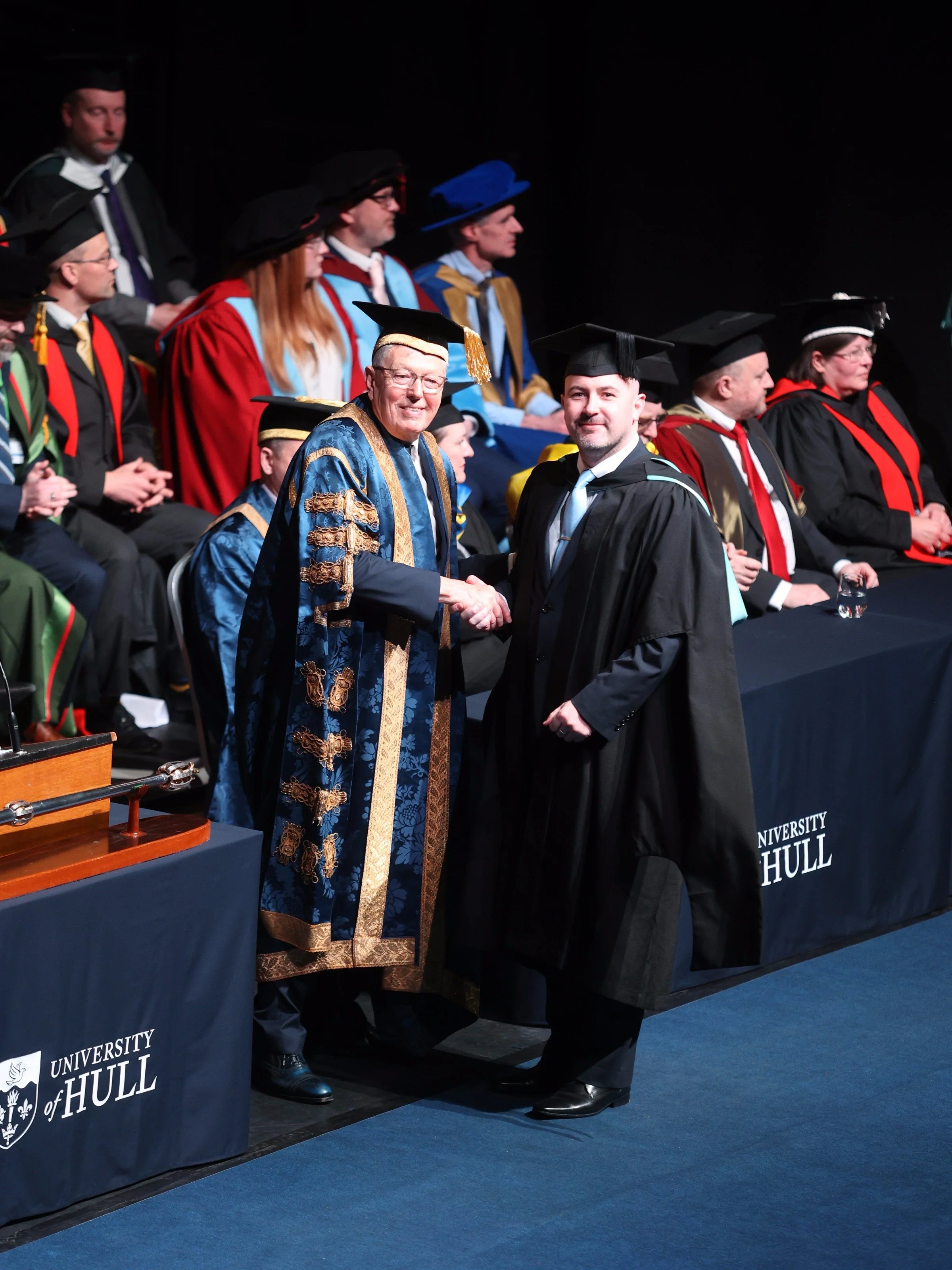 University graduation ceremony at University of Hull featuring the founder of BSA in cap and gown shaking hands with a university official in academic regalia, with other officials and faculty members seated on stage.