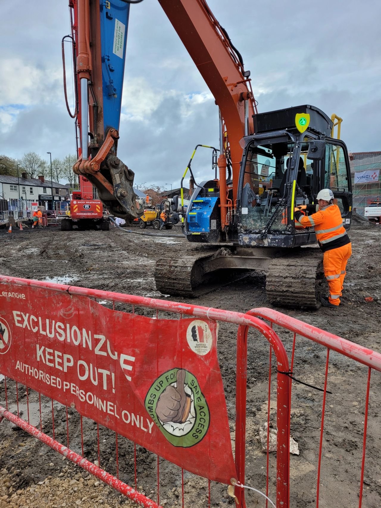 Construction site with a worker in orange uniform and white helmet operating an excavator, surrounded by safety barriers and signs.