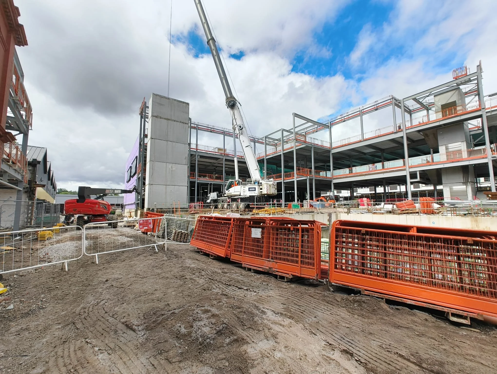 Construction site with a crane, orange safety barriers, and a partially built multi-story building under a partly cloudy sky.