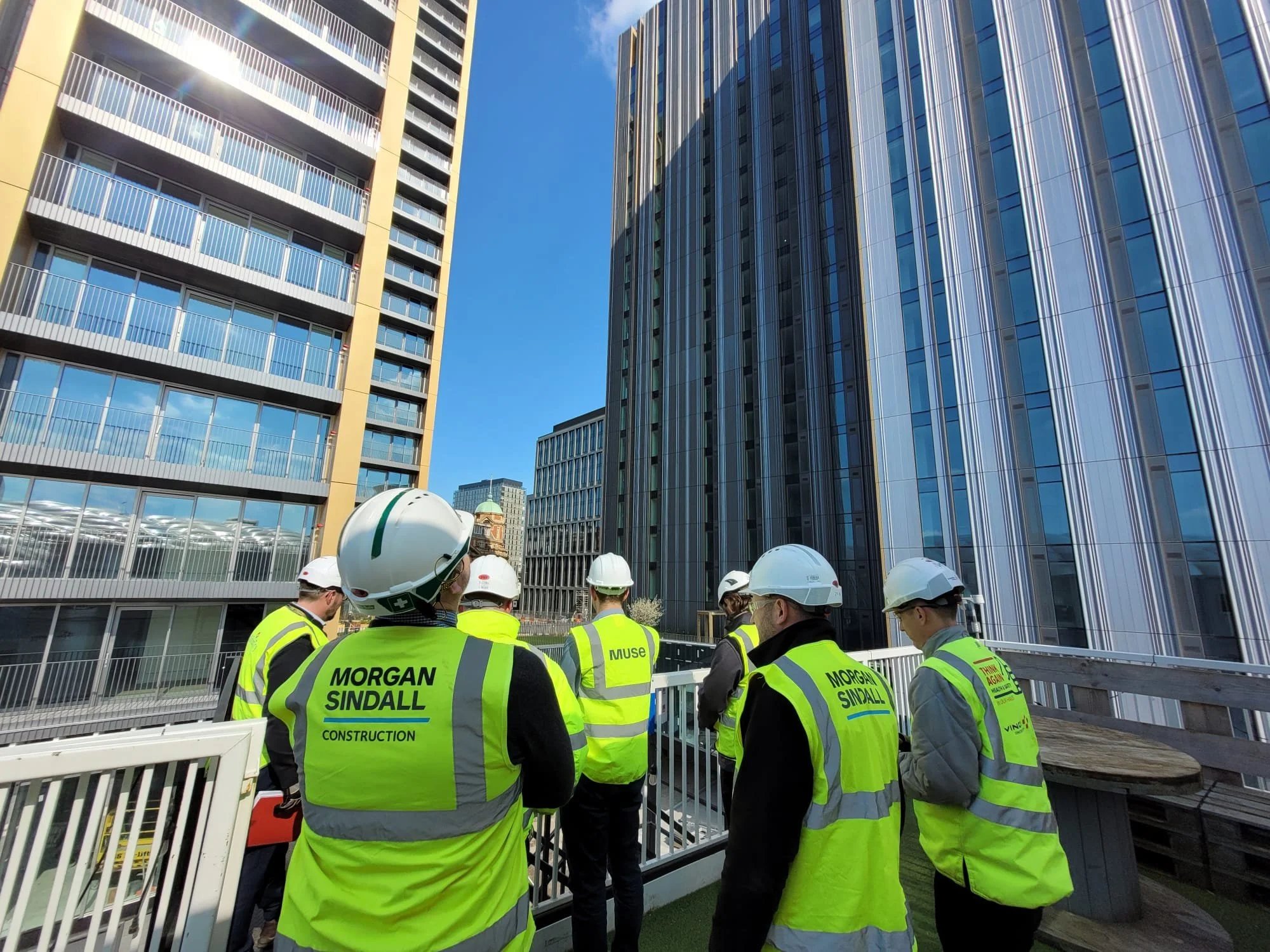 Group of construction workers wearing yellow safety vests and white helmets standing on a balcony, surrounded by tall modern glass buildings in an urban setting.