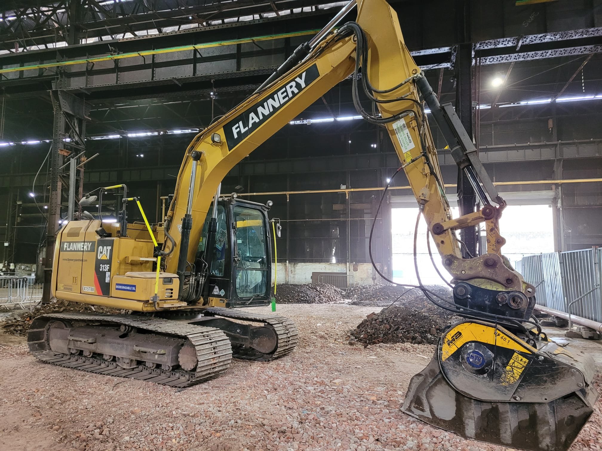 A large yellow excavator with black tracks inside a warehouse, digging into dirt and gravel.