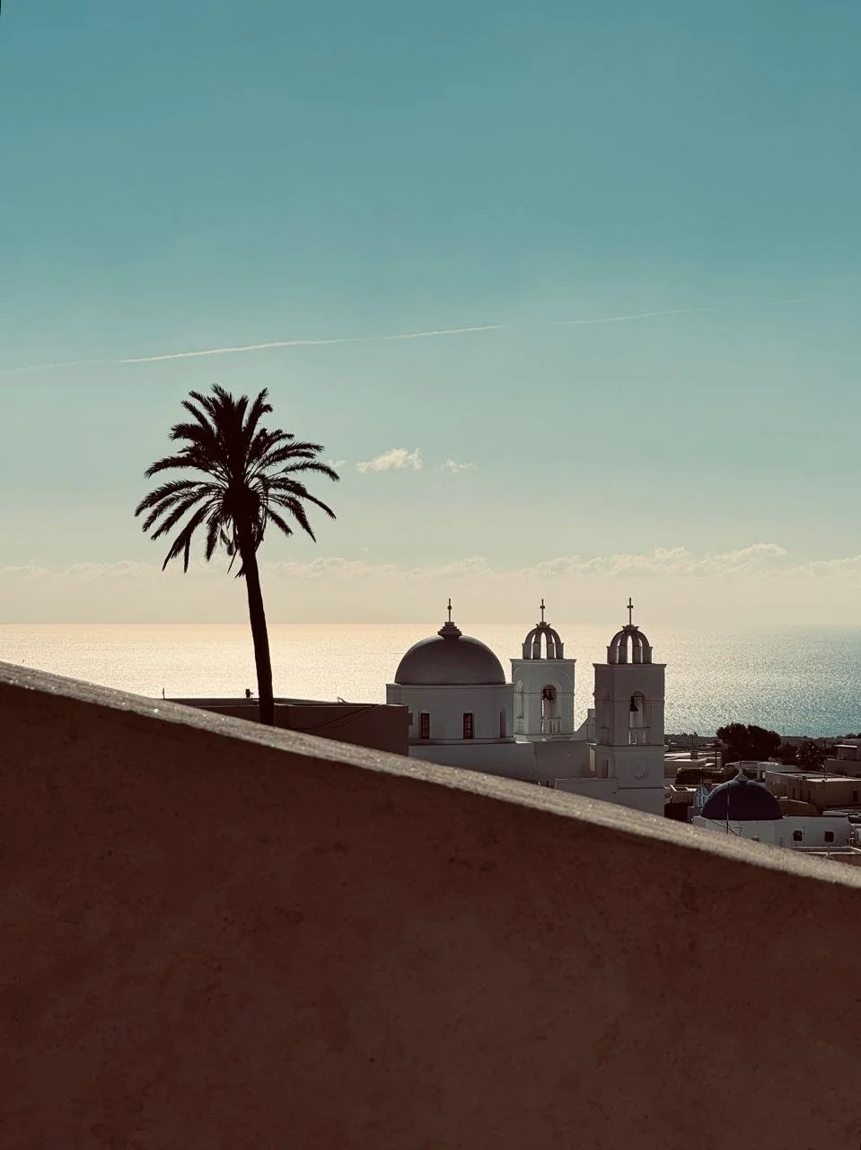 Scenic view of a white church with a dome and two bell towers, a tall palm tree in the foreground, and the ocean in the background with a clear sky in a minmal cycladic scenery in Santorini.