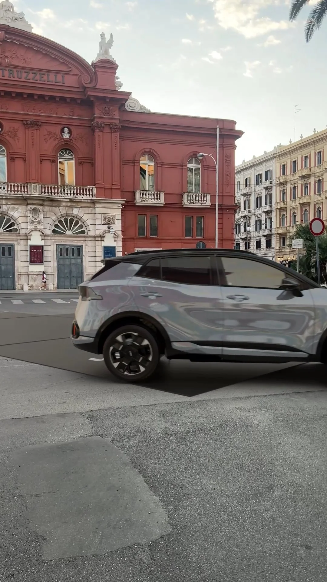 Un'auto moderna di colore grigio che attraversa una strada cittadina con edifici storici sullo sfondo.