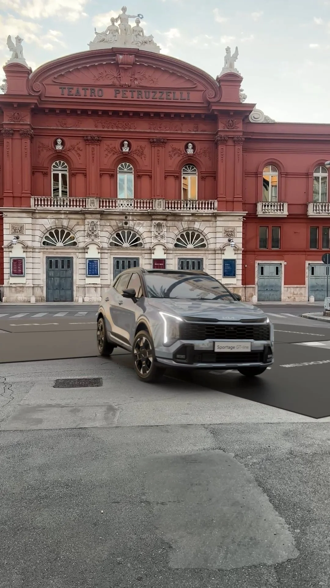 Un'auto grigia parcheggiata davanti a un edificio storico di colore rosso con colonne e statue in cima, con il nome 'Teatro Petruzzelli'.