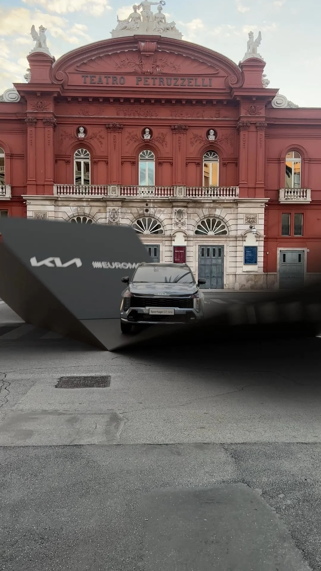 Vettura Kia Sportage GT-line davanti al Teatro Petruzzelli di Bari, visibile molteplici dettagli architettonici e sculture sulla facciata, con un cartellone pubblicitario in primo piano