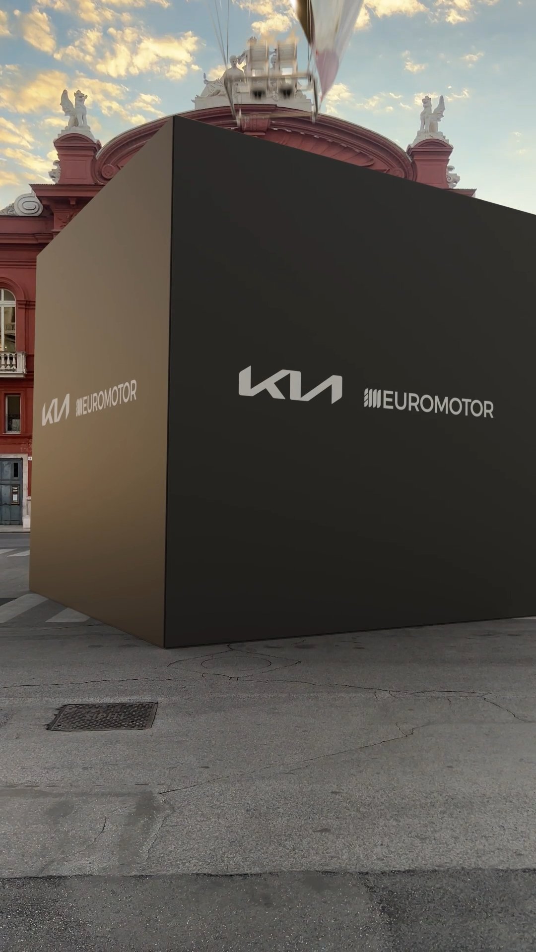 Una grande scatola nera con i loghi della Kia e Euromotor, posizionata su una strada asfaltata, con un palazzo storico in stile Barocco con statue sul tetto sullo sfondo e il cielo al tramonto.