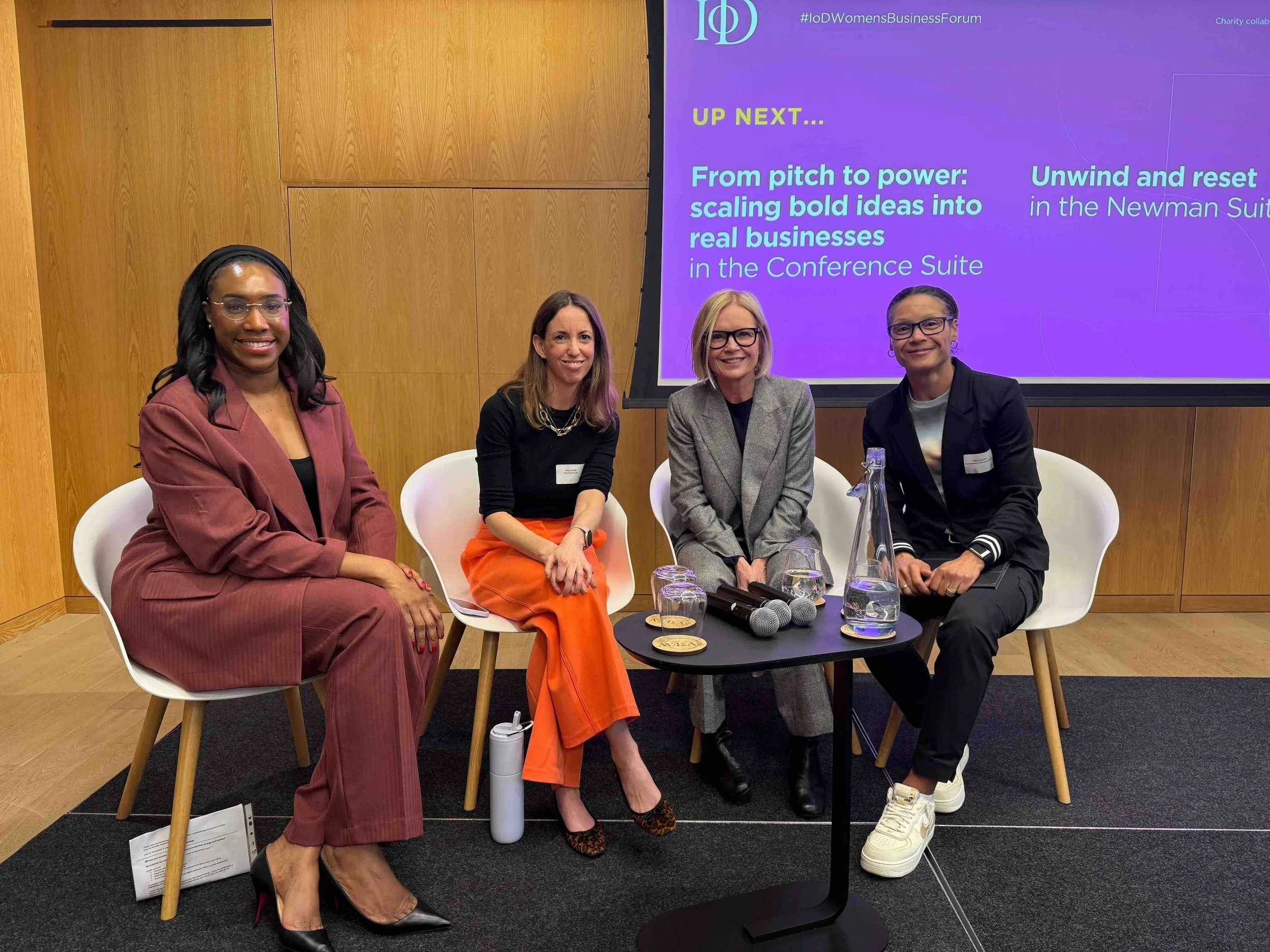 Four women seated on chairs in front of a purple presentation screen at a conference. The women are smiling and dressed in business attire. The table in front of them has two microphones, a water pitcher, and glasses. The presentation screen displays event information and schedule.