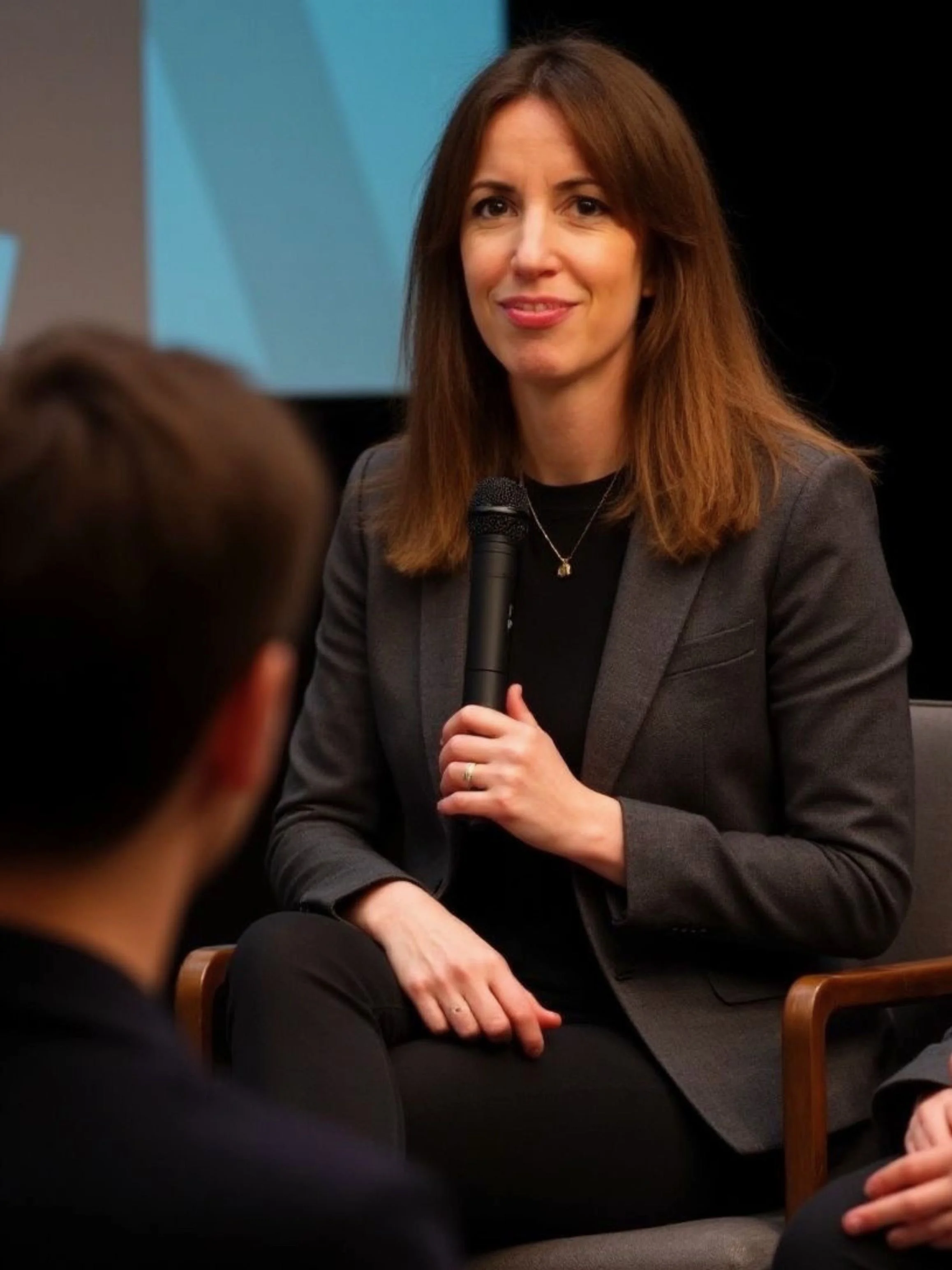 A woman with long brown hair, wearing a gray blazer and black top, sitting and holding a microphone during a discussion or interview.