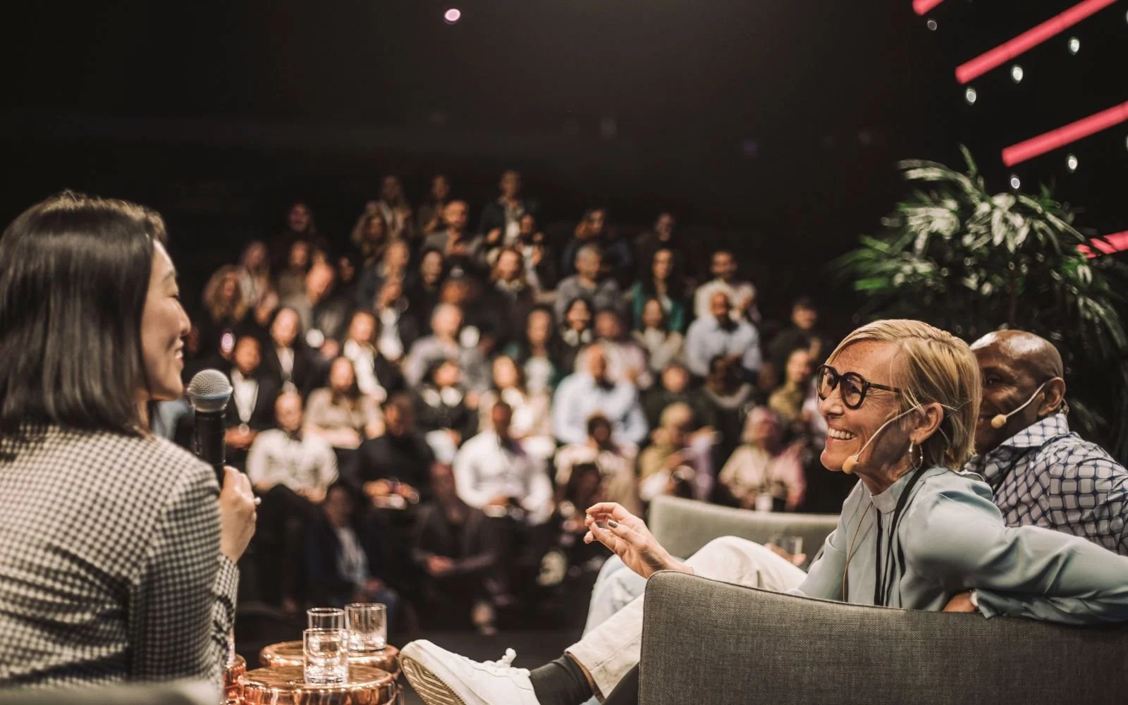 A woman holding a microphone and speaking to an audience, smiling. Two seated persons are in the foreground, smiling and listening. The audience is in the background, blurred.
