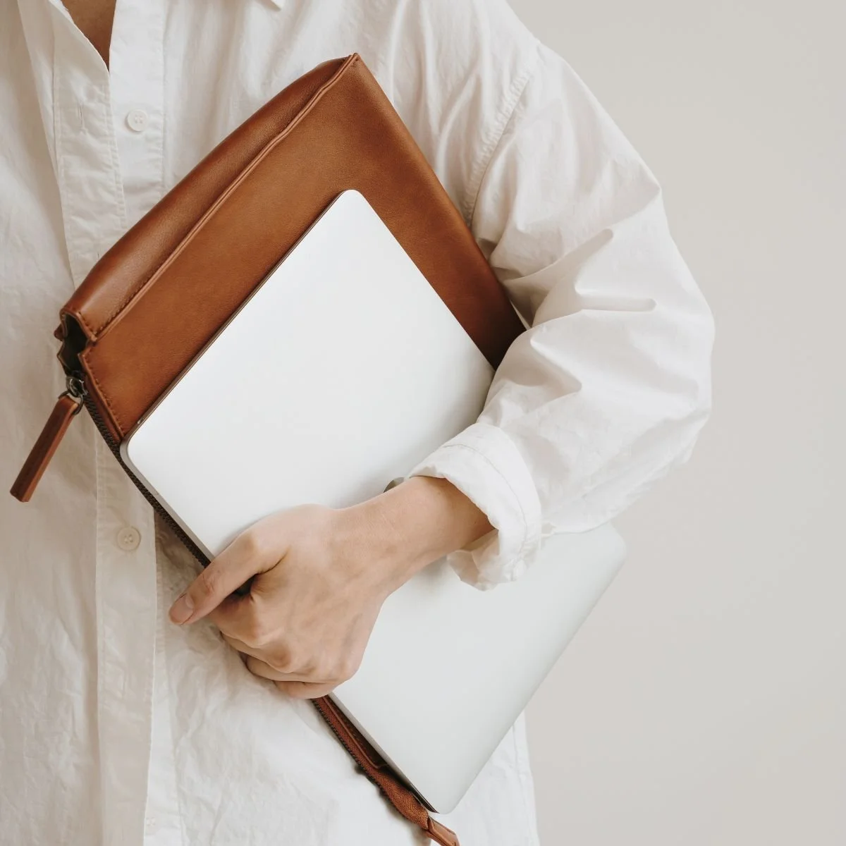 Person in a white shirt holding a laptop and a brown leather pencil case or pouch.