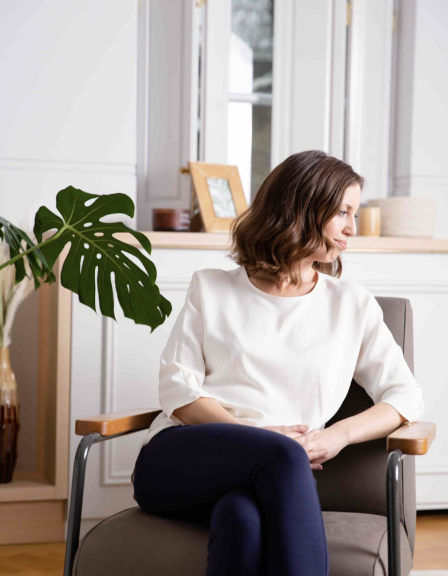 A woman with brown wavy hair sitting on a chair in a bright room, wearing a white top and dark pants, with a large green plant and framed photo in the background.