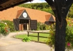 A rustic building with a large clock on the front, surrounded by greenery and a gravel pathway, with a tree in the foreground.