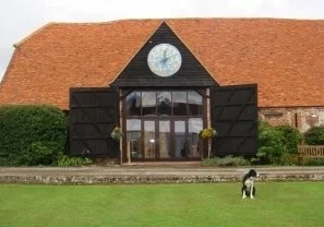 A building with a large clock on top and black barn-style doors open in the front. There is a grassy area in front with a dog walking on it, and bushes are visible to the sides of the building.