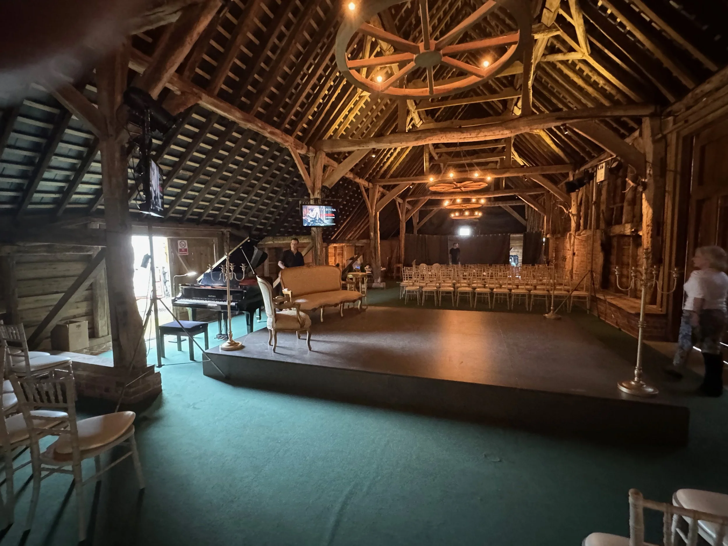 Empty rustic barn with a small stage setup featuring a grand piano, vintage furniture, and rows of chairs, illuminated by warm lighting, ready for a performance or wedding.