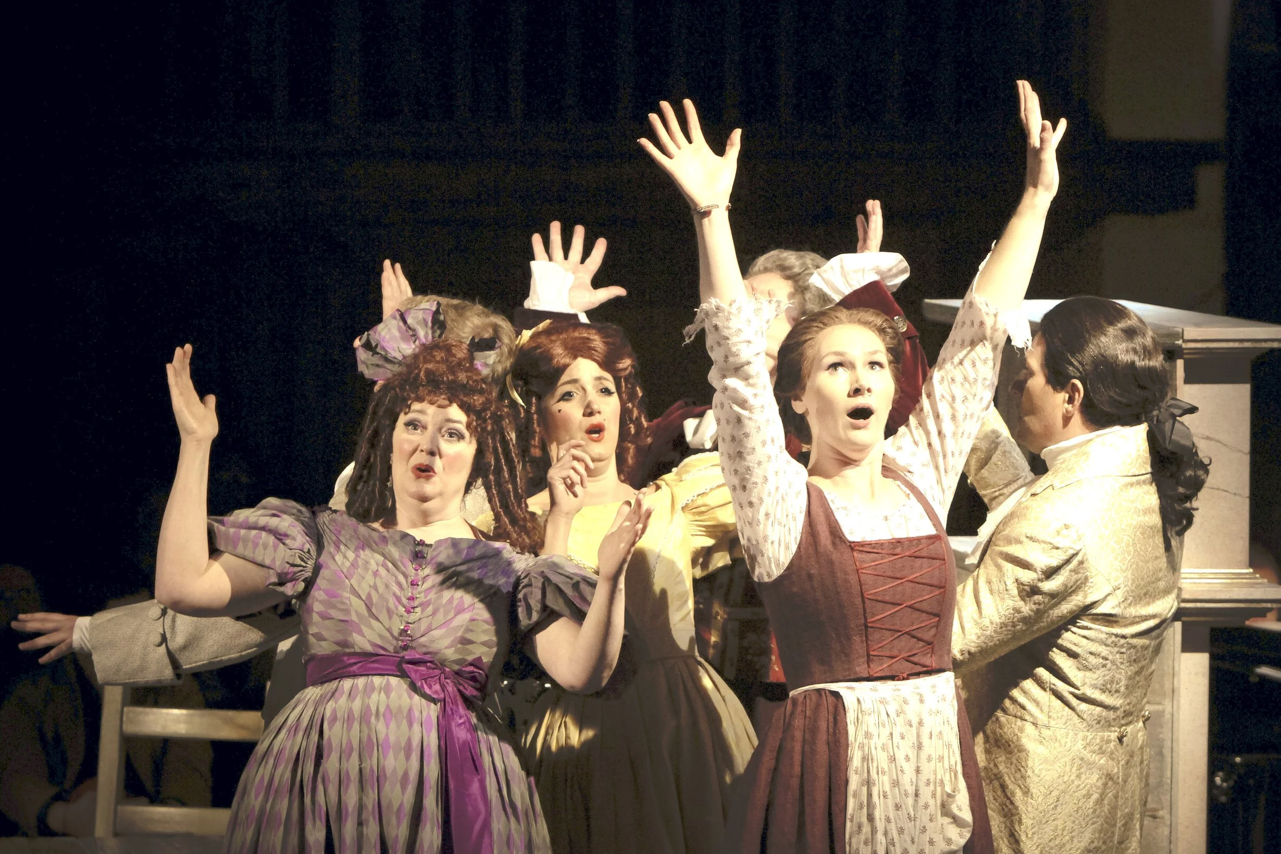 Actors on stage dressed in historical costumes, performing with expressive facial reactions and raised hands.