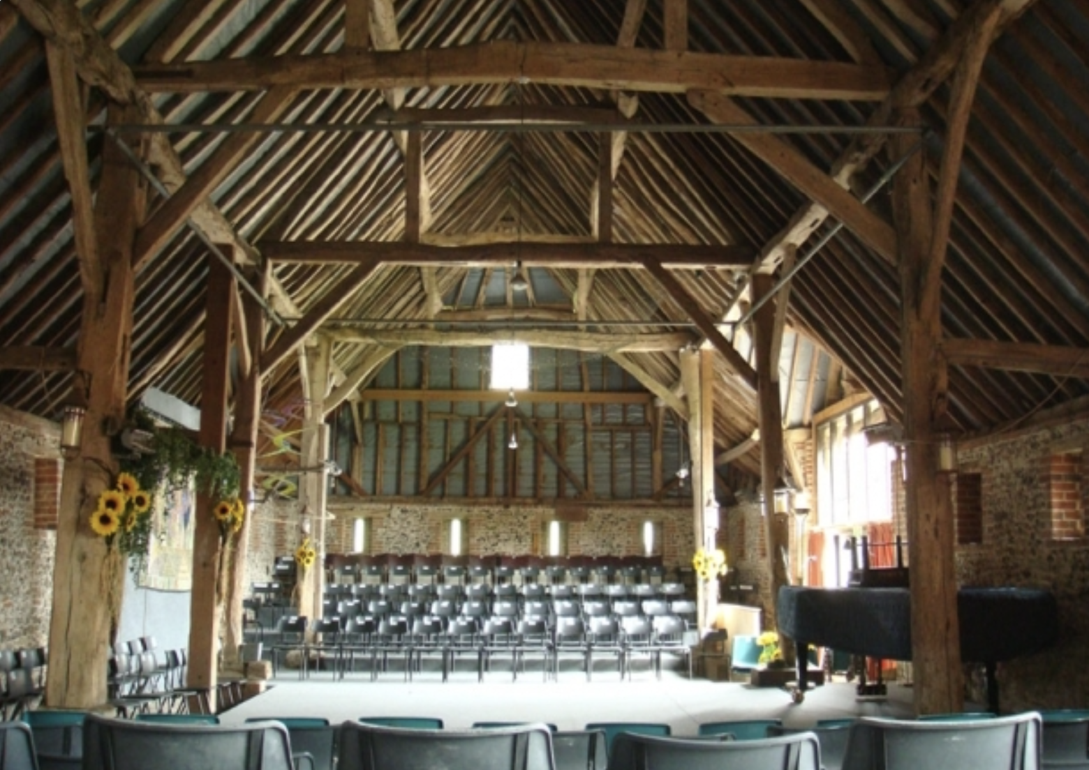 Interior of a rustic barn with wooden beams, brick walls, and stage area with chairs and a grand piano, decorated with sunflowers and hanging lights.