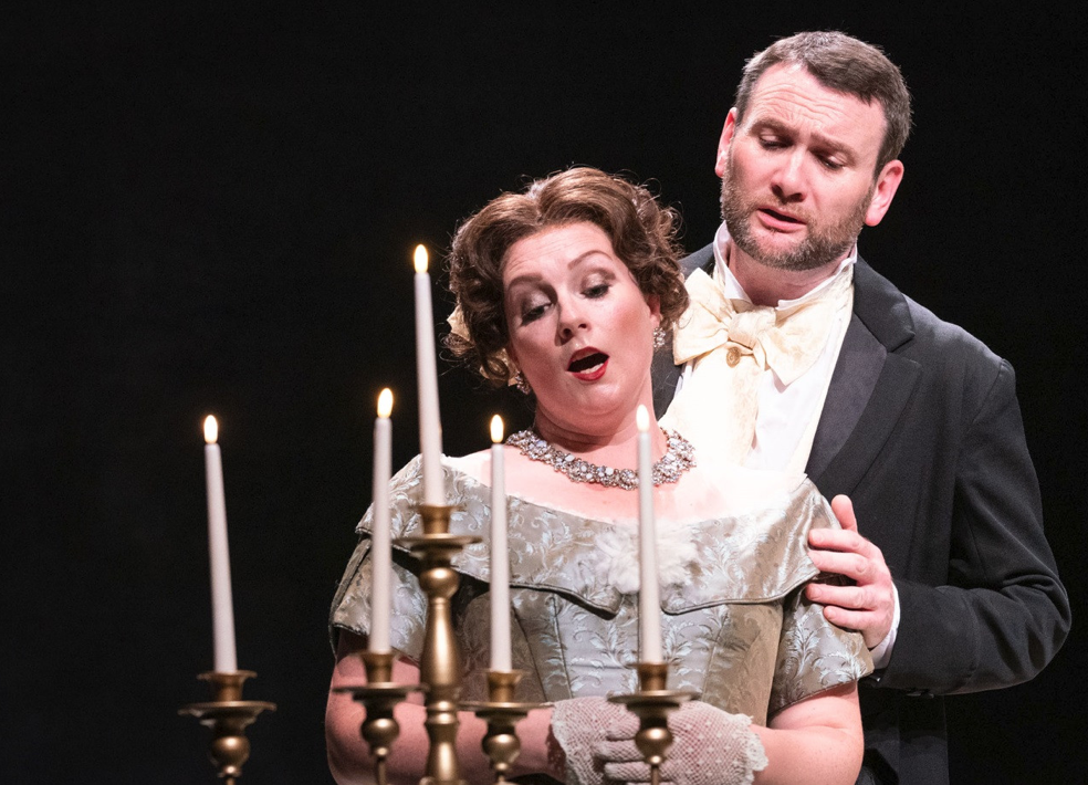 A theatrical scene with a woman in an elegant dress and necklace, and a man in a tuxedo, standing close to a candlestick holder with lit candles against a black background.