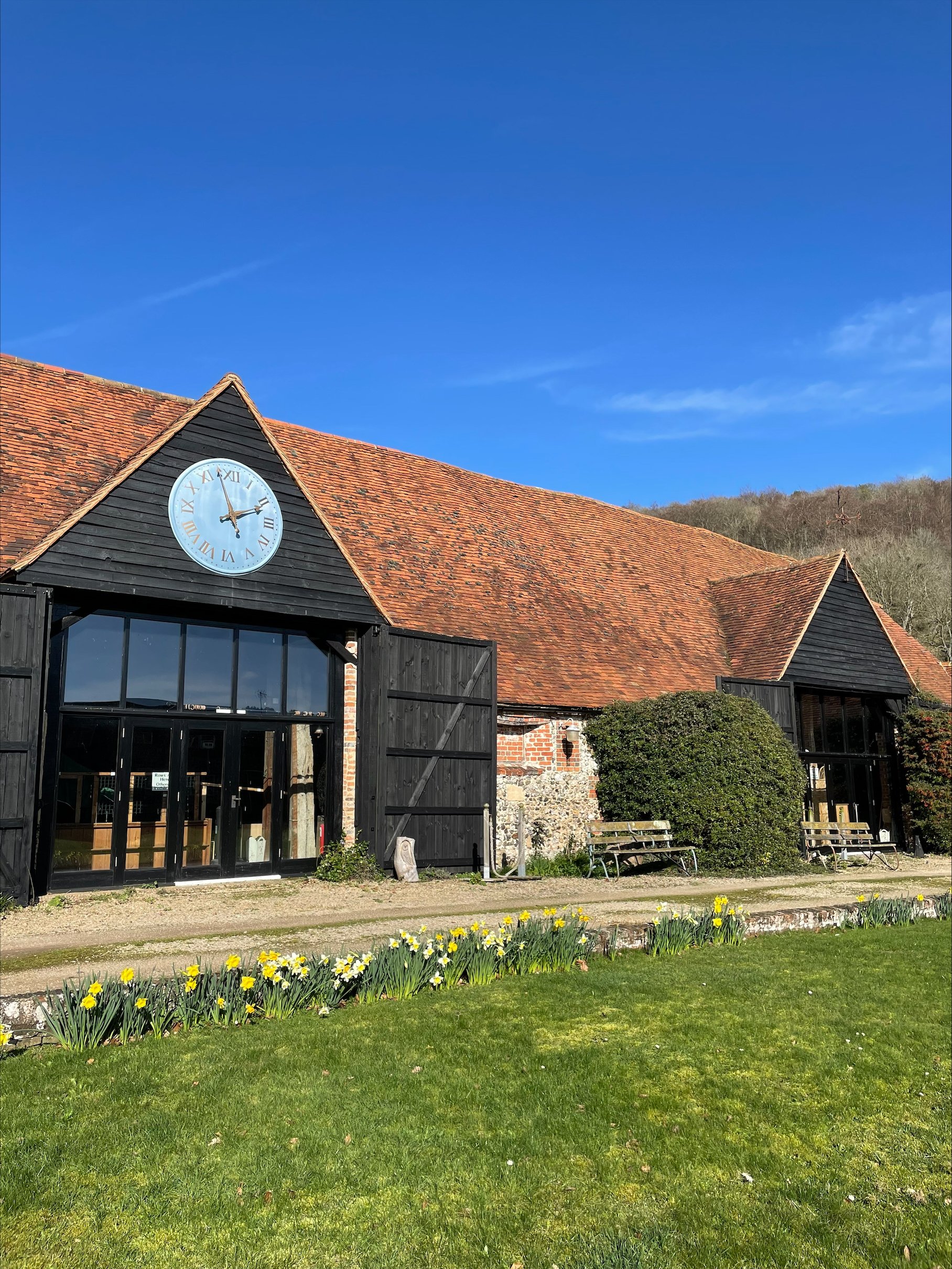 A rustic barn with a large clock on the gable, black wooden doors, and a garden with yellow flowers in front, under a clear blue sky.