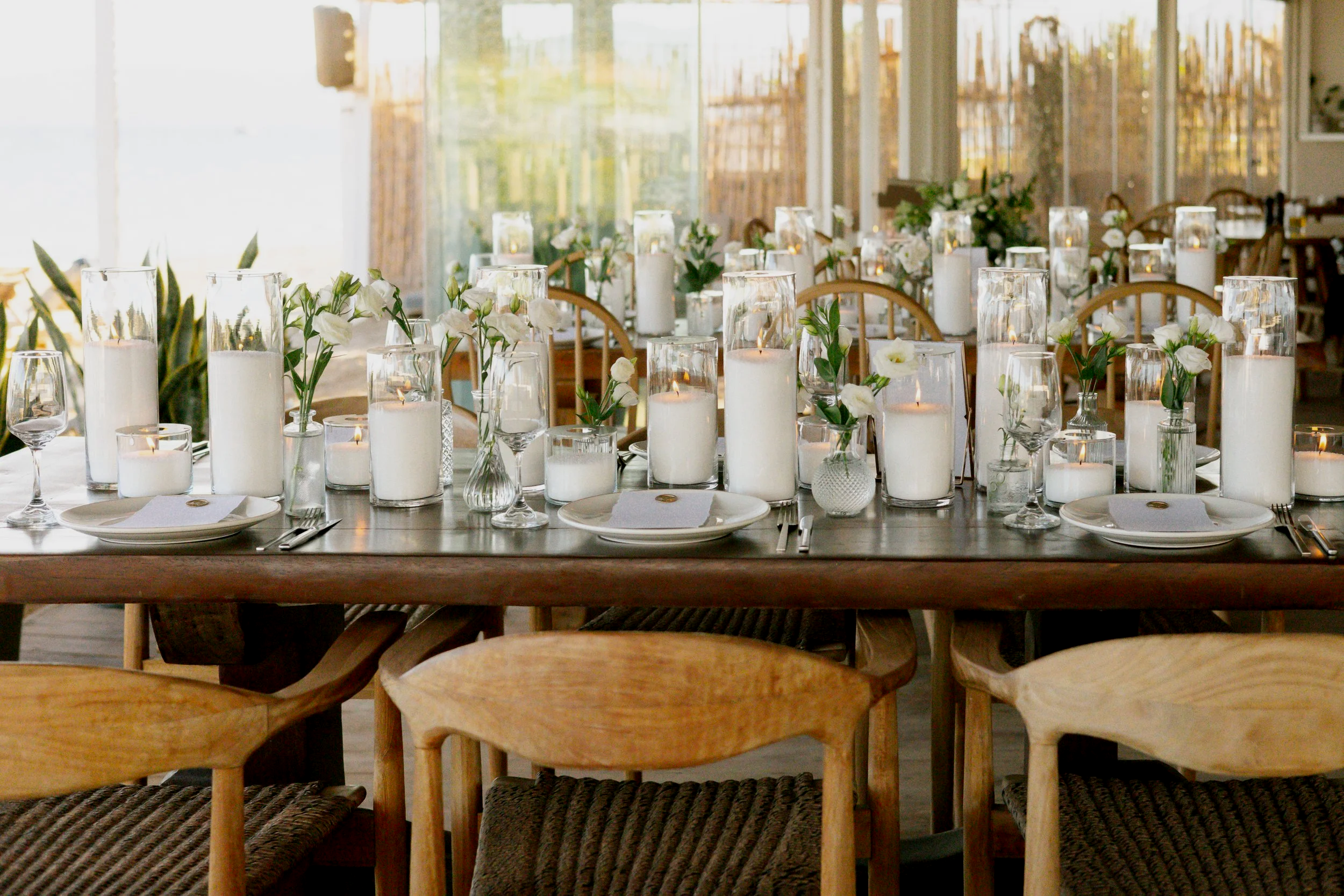Table de mariage décorée avec des bougies blanches dans des vases en verre et des fleurs blanches, dans une salle lumineuse avec de grandes fenêtres.