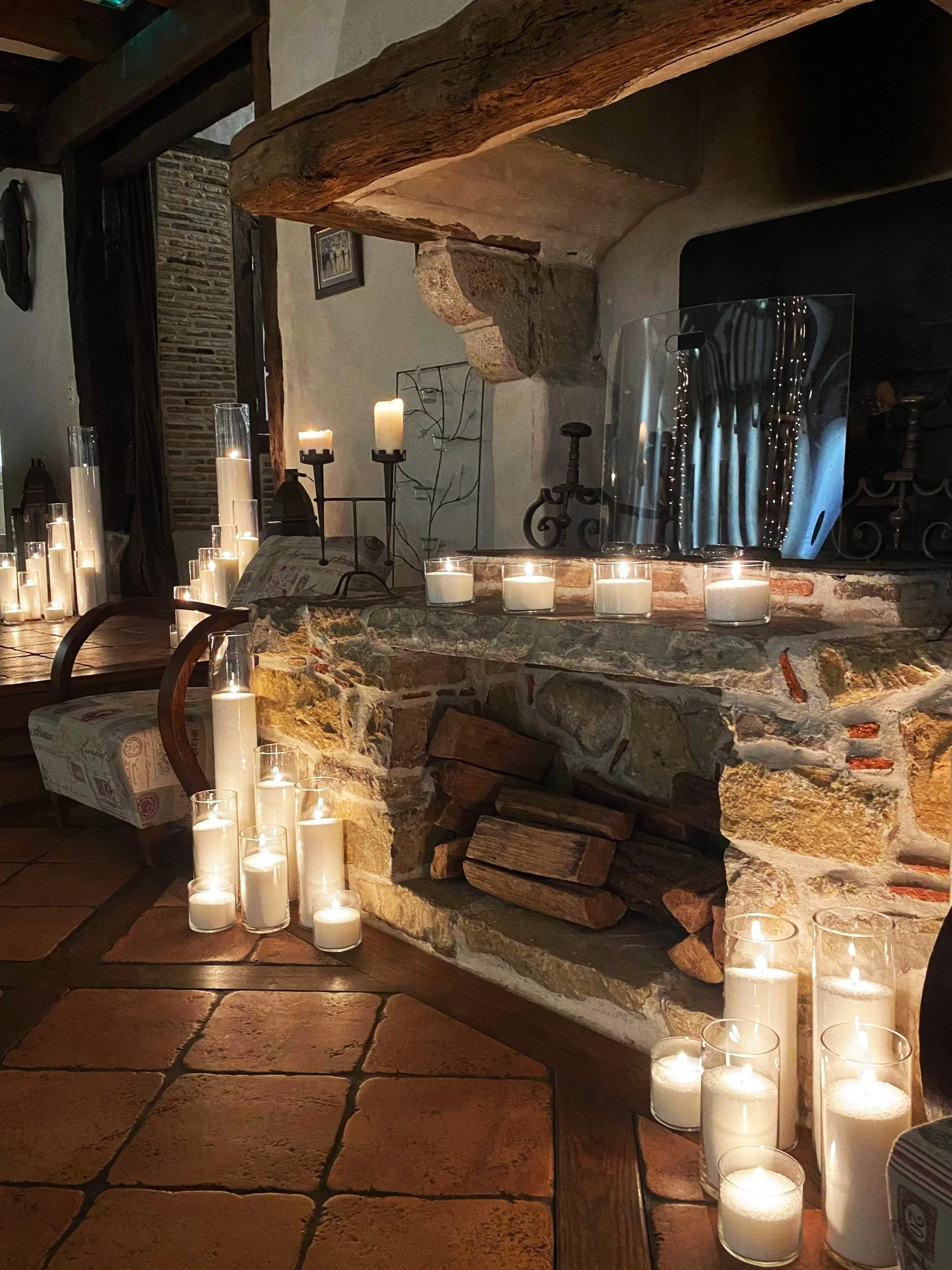 Une cheminée en pierre allumée entourée de nombreuses bougies dans des votives en verre, dans une ambiance chaleureuse et rustique.