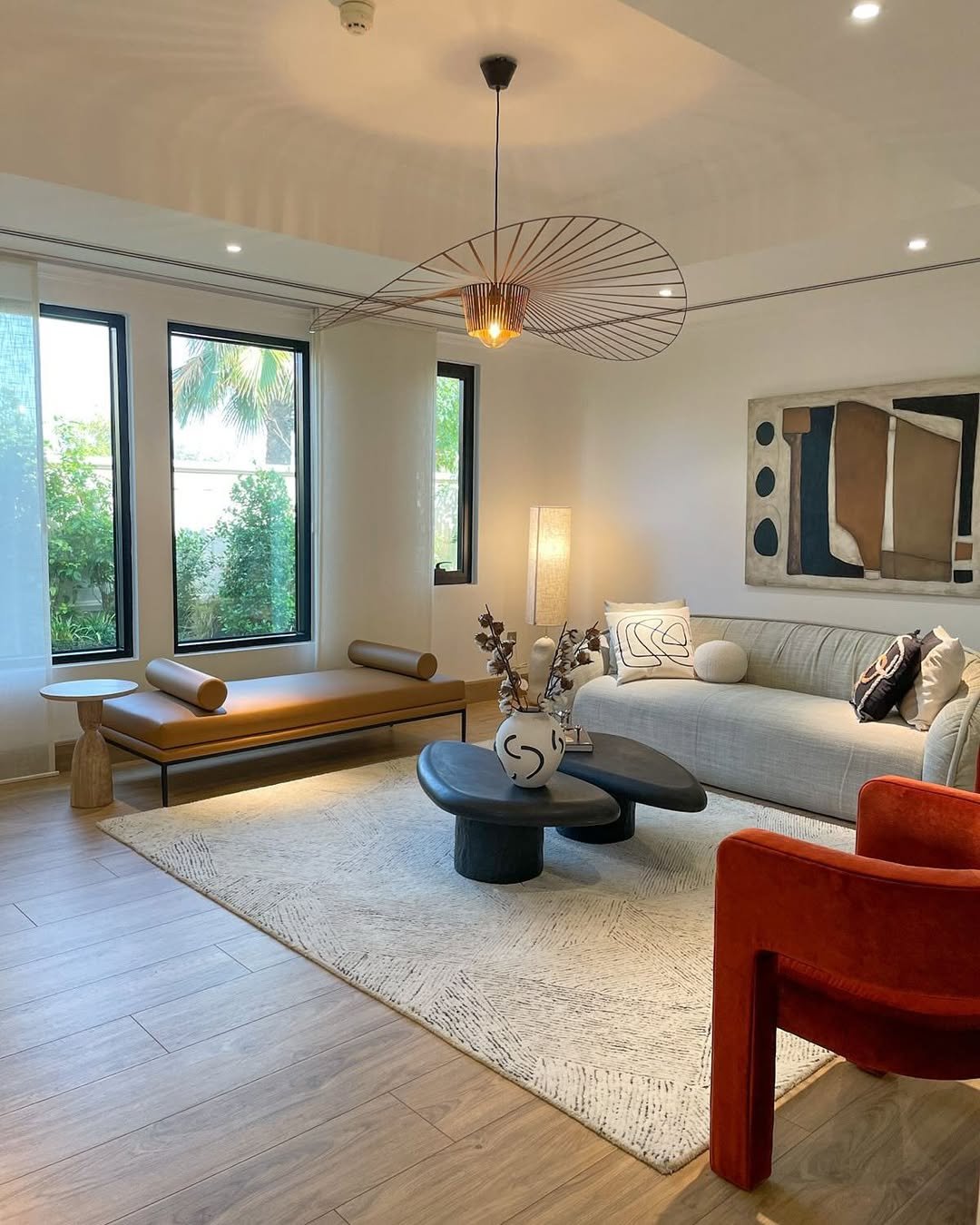 Rasha Ghazaleh | Modern living room with large windows, contemporary furniture, a white couch with pillows, a black coffee table with vases, a leather bench by the windows, a red armchair, a textured rug, wall art, and a ceiling lamp.