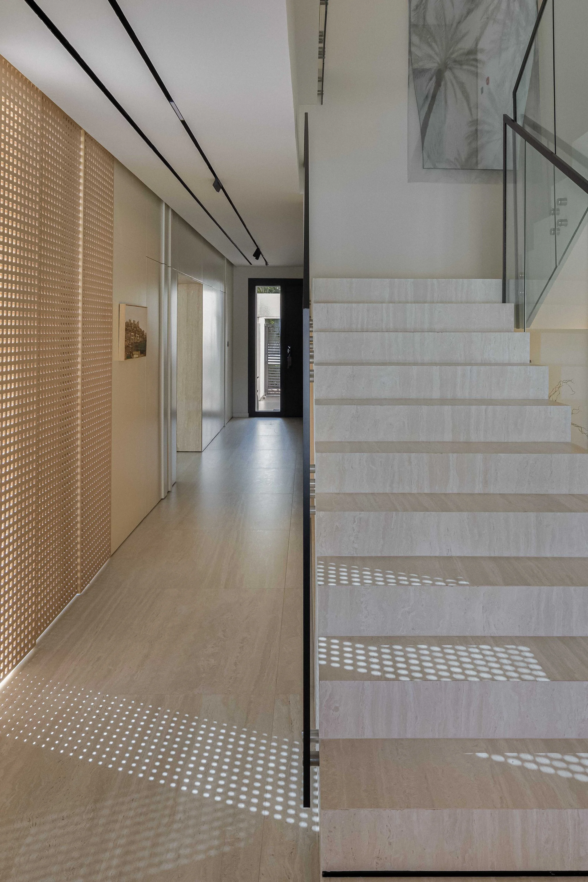 Modern interior hallway with beige walls and a staircase on the right side with glass railing, through which shadow patterns are cast on the floor from the sunlight shining through the perforated wall on the left.