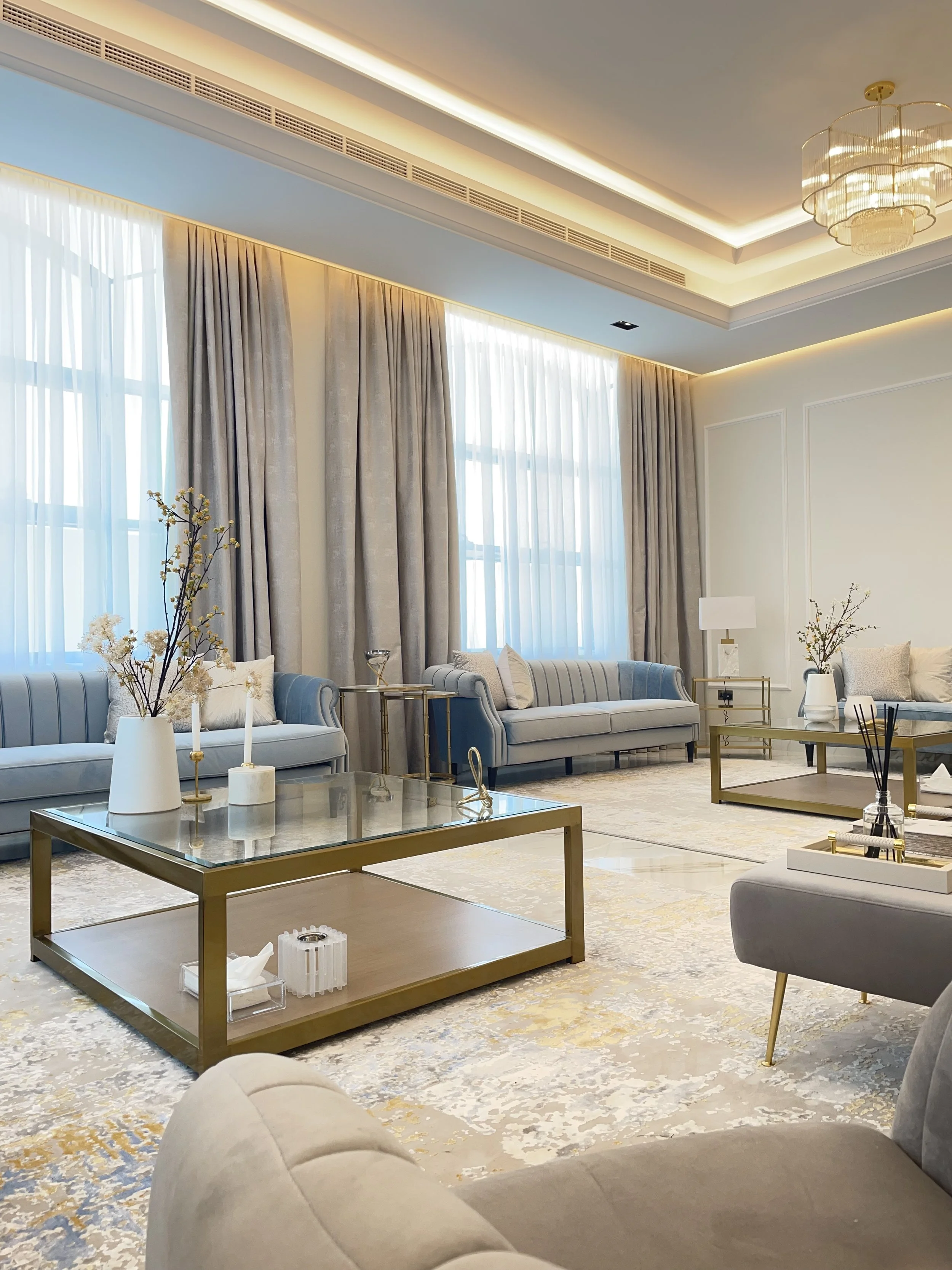 Elegant living room with large windows, beige and blue sofas, gold coffee tables, decorative vases, and a beige area rug.