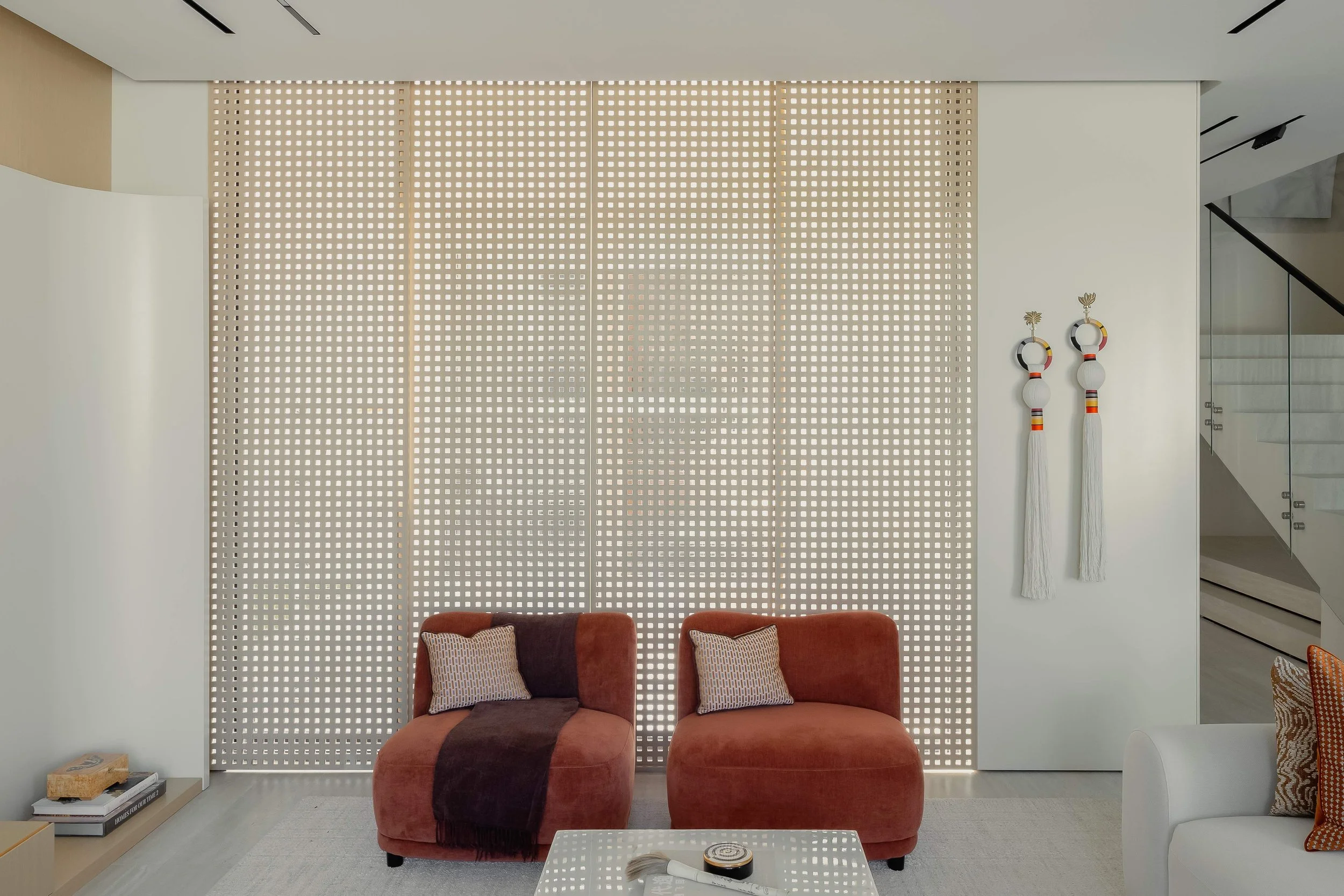 Living room with two orange armchairs, two pillows, a white coffee table with decorative items, a white wall with two hanging art pieces, and a staircase in the background.