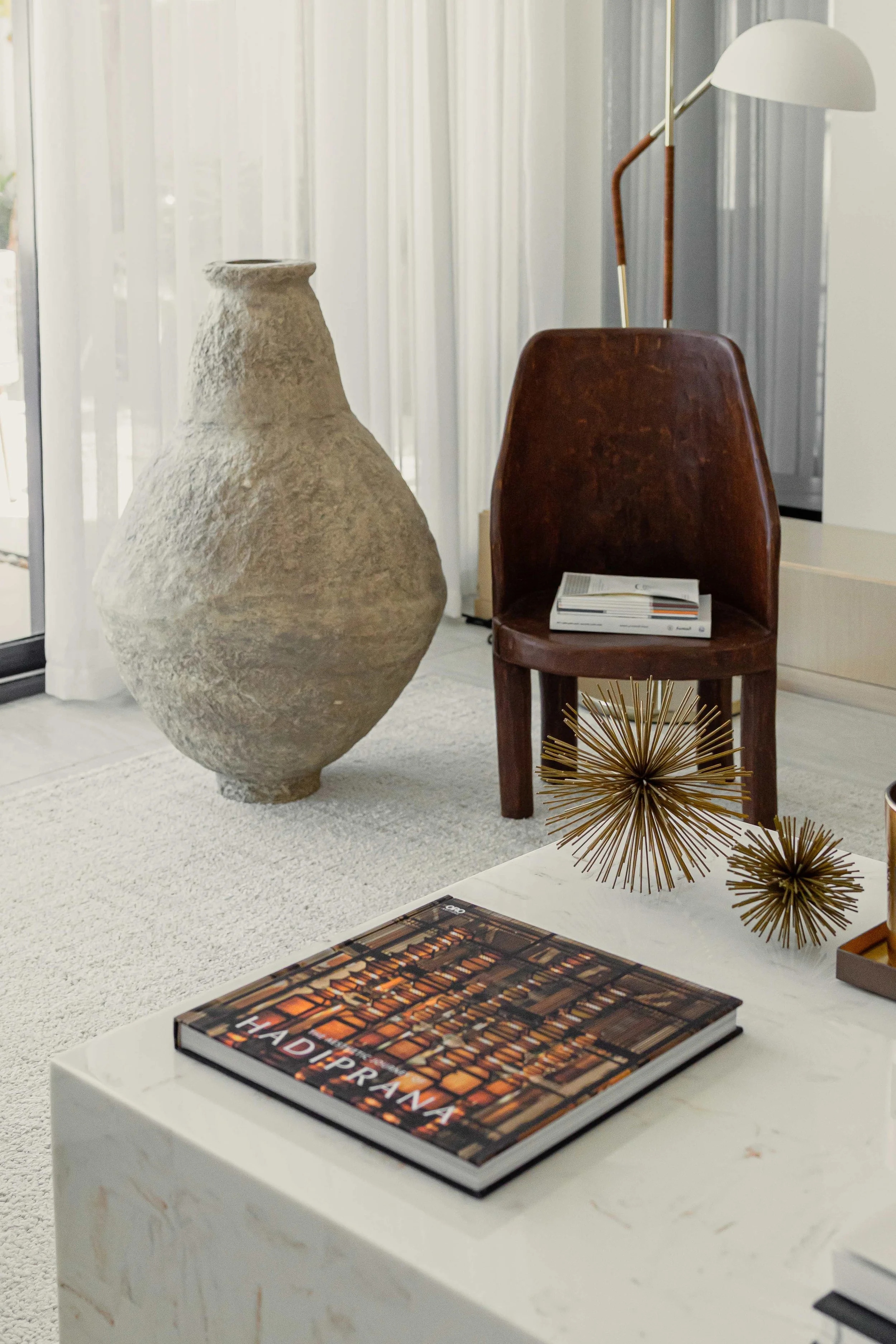 Living room corner with large textured vase, wooden chair with magazines, gold geometric decor, and a white table with a book titled 'HAD PRANA'.