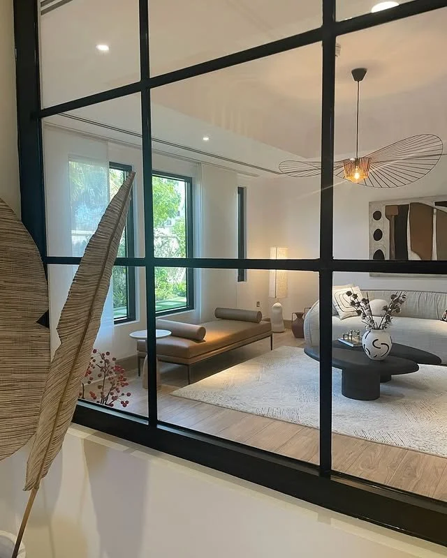 Modern living room seen through a large black-framed glass partition. It features a geometric light fixture, a beige bench with cylindrical cushions, a white area rug, a round black coffee table with a vase, a bed with pillows, and large windows with greenery outside.