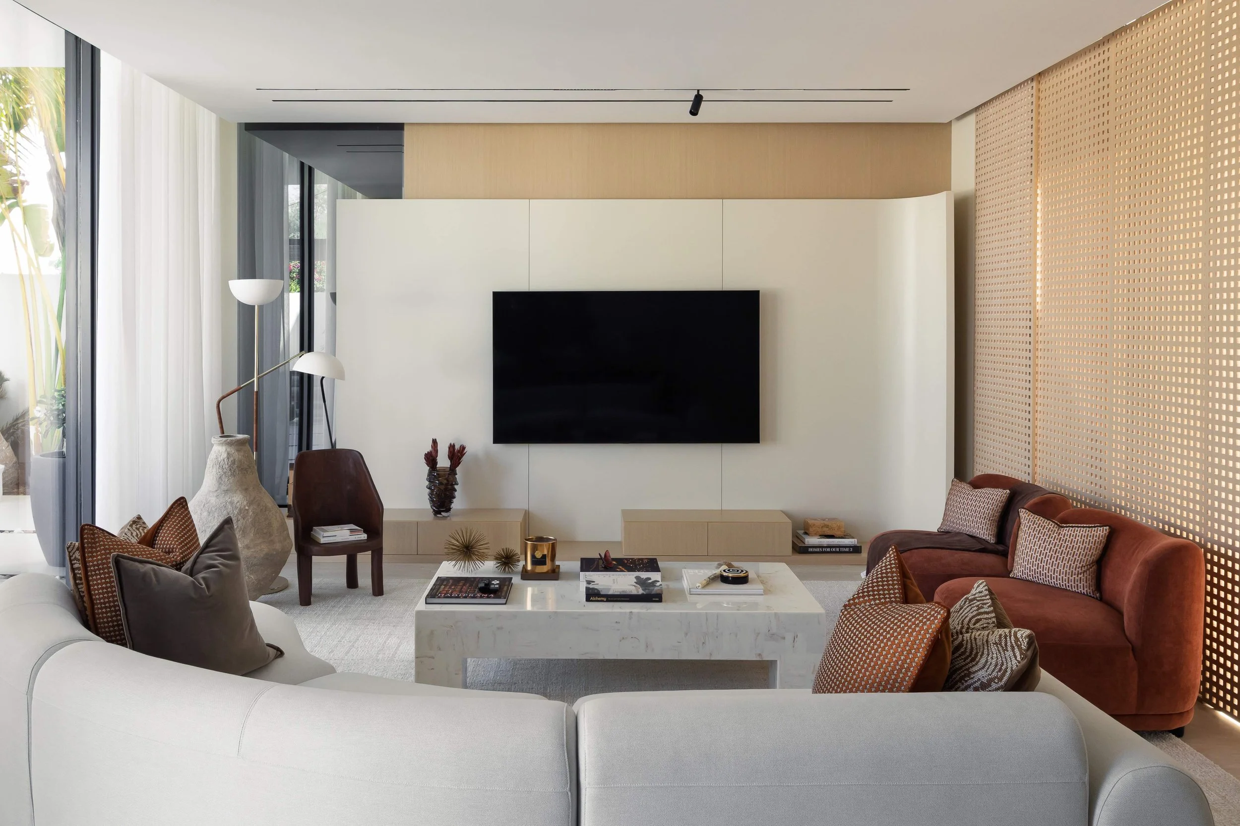 Modern living room with white sectional sofa, red velvet chairs, a marble coffee table, wall-mounted TV, decorative pillows, side tables, and large windows with curtains.